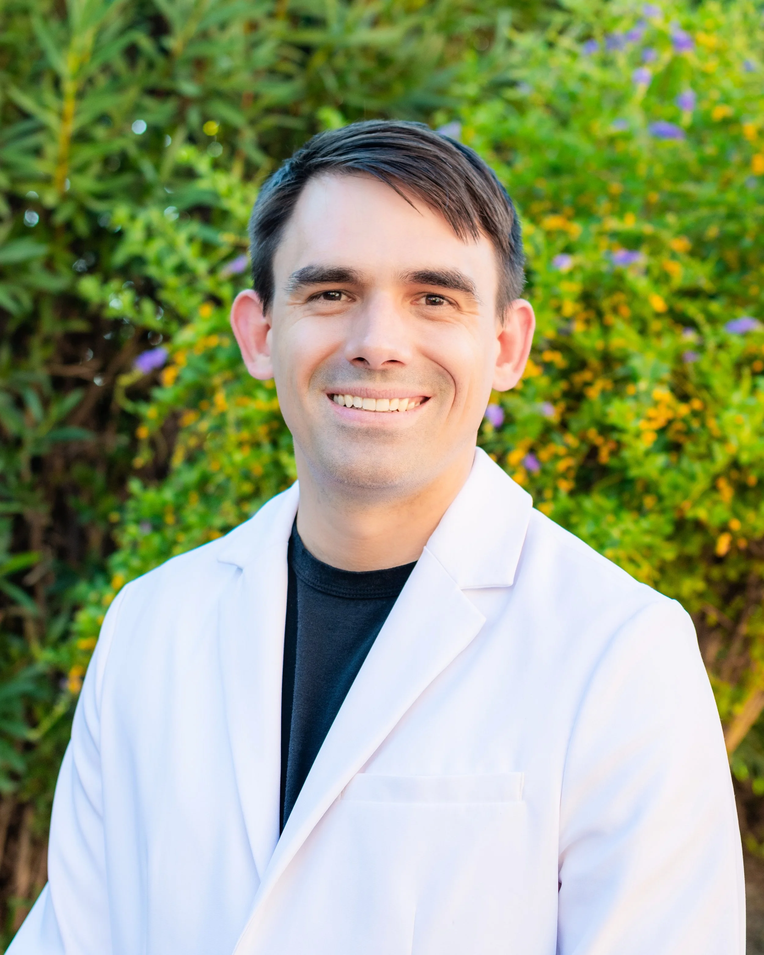 Photo of Dr. Sean McEvoy. Close-up of a smiling man with dark hair, wearing a white coat over a black shirt, standing outdoors with green bushes and purple flowers in the background.
