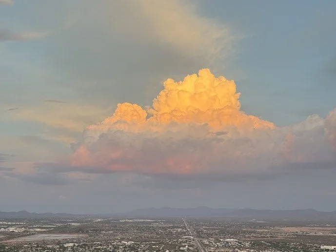 A vast landscape with a large, colorful cloud illuminated by the setting or rising sun, with a distant horizon and mountains in the background.