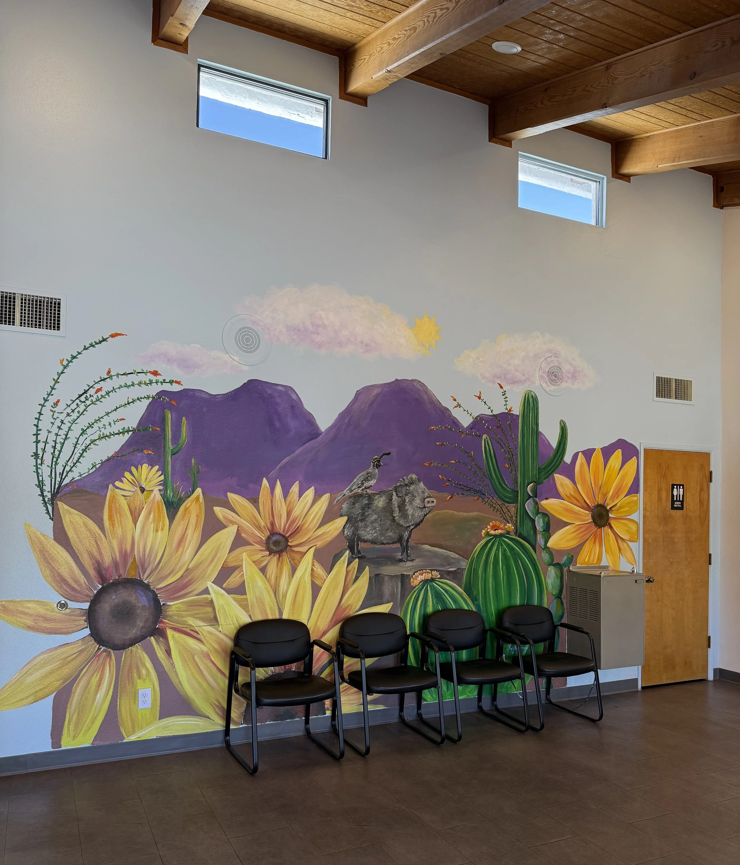 El Sol Office, the interior of a room with a wall mural of a desert scene featuring mountains, cacti, flowers, a bird, and a sun. Four black chairs are lined up against the mural, and there are windows and ceiling lights visible.