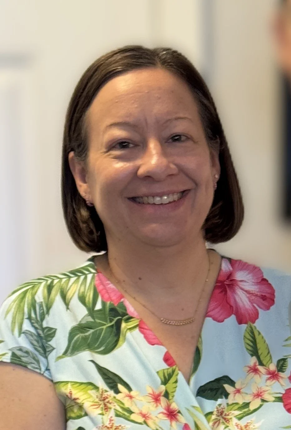 Photo of Dr. Narissa Whitelaw. Smiling woman with short brown hair wearing a floral shirt with pink and green patterns, gold necklace, and earrings indoors.