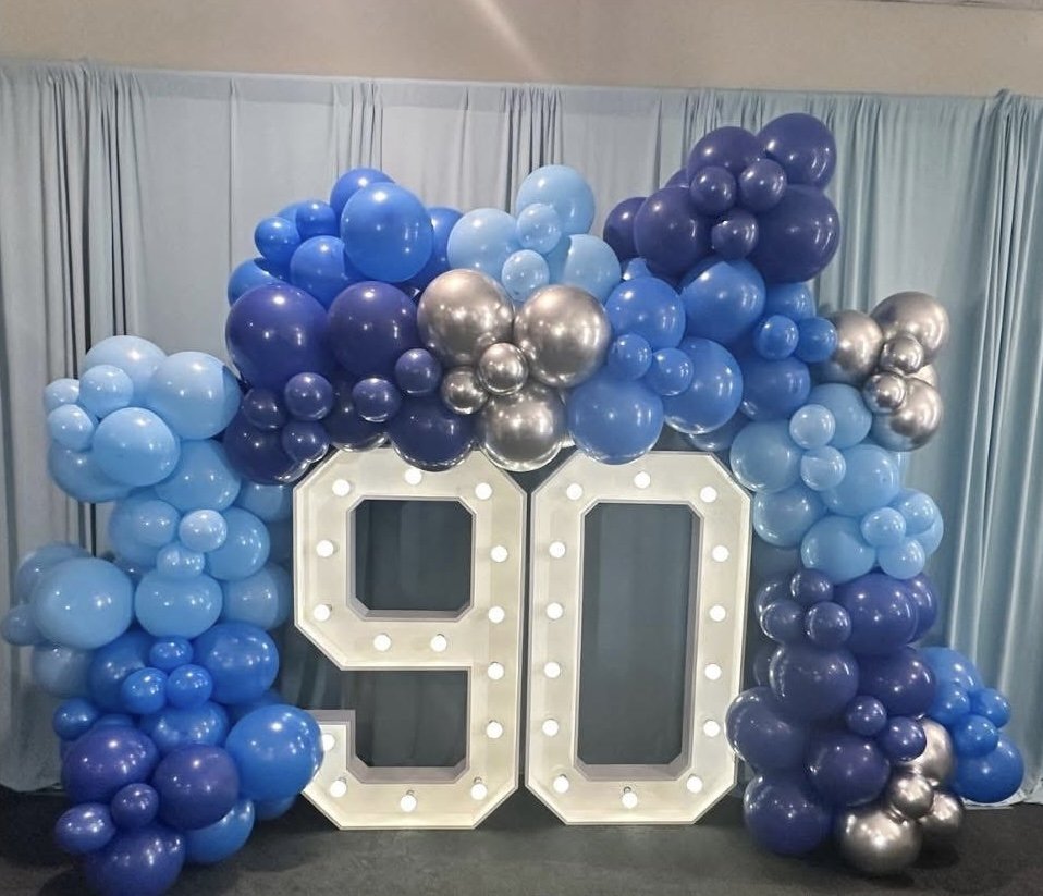 Large lighted number 90 decorated with blue, purple, and silver balloons in a celebration setting.