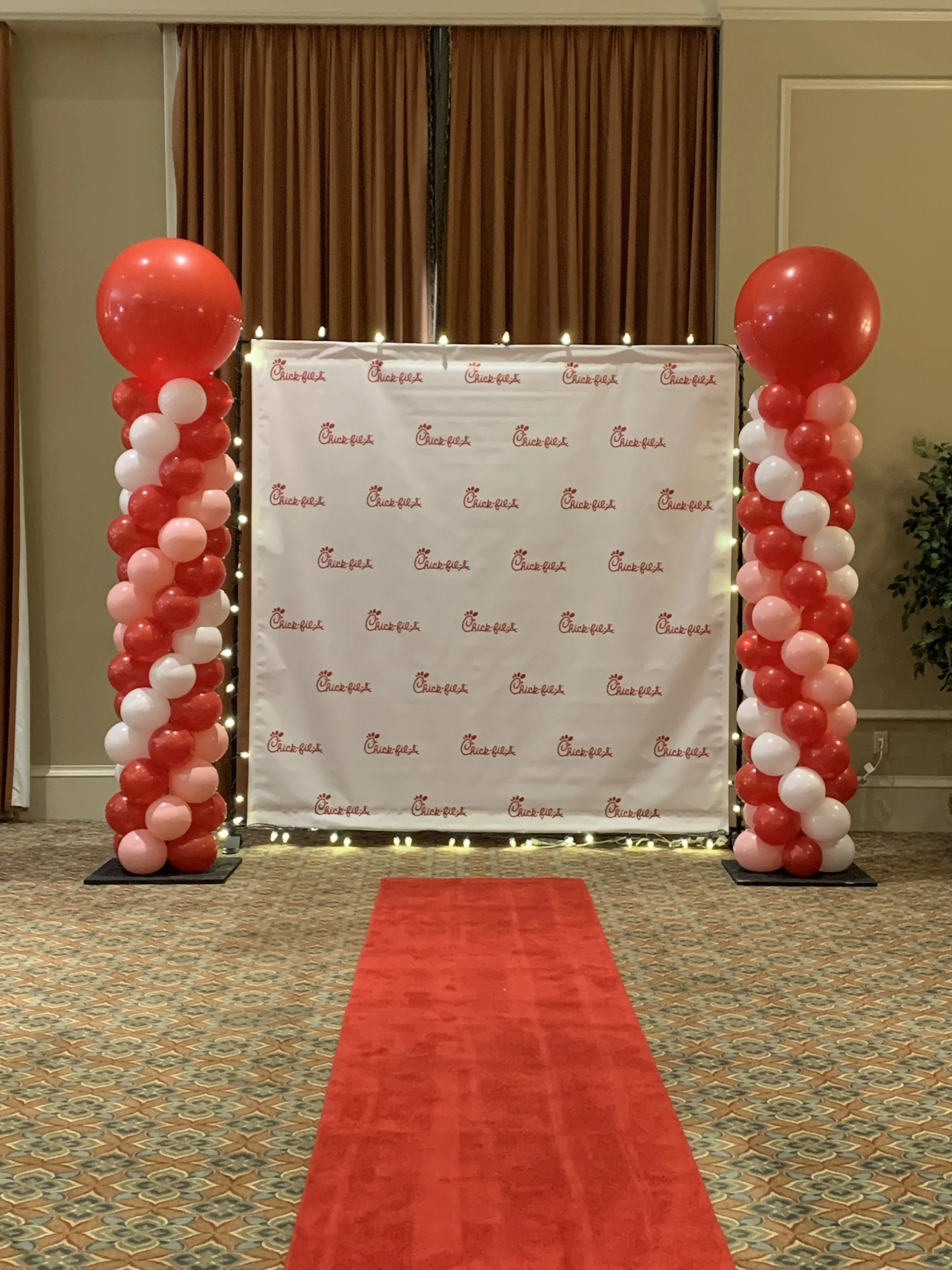 Indoor event setup with a red carpet leading to a backdrop covered in Chick-fil-A logos, flanked by balloon columns of red, pink, and white balloons, and illuminated string lights.