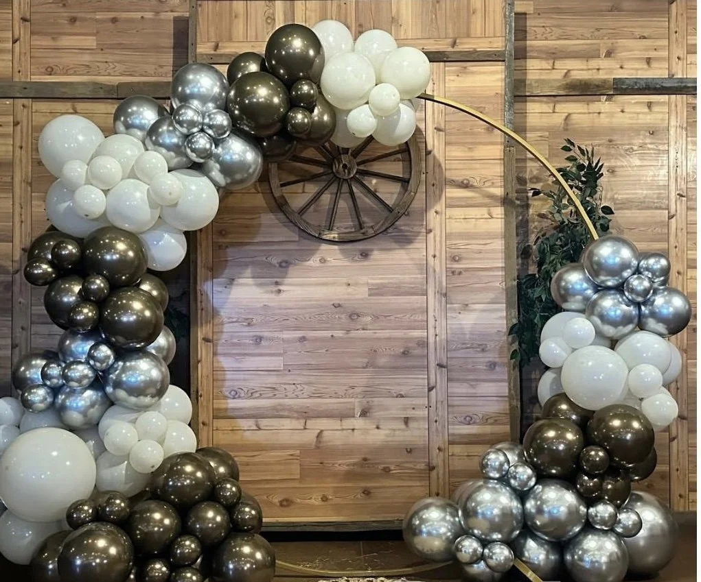 Decorative balloon arch with black, white, and silver balloons against a wooden wall with a wagon wheel decoration and a green plant.