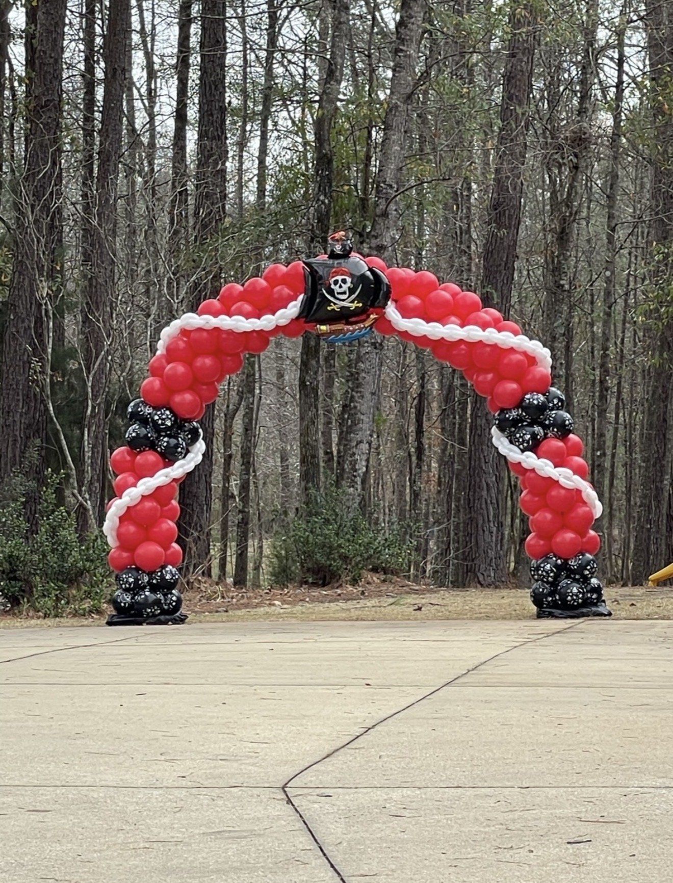 A decorative arch made of red, white, and black balloons, with a pirate-themed balloon decoration at the top, set outdoors in a wooded area with trees in the background.