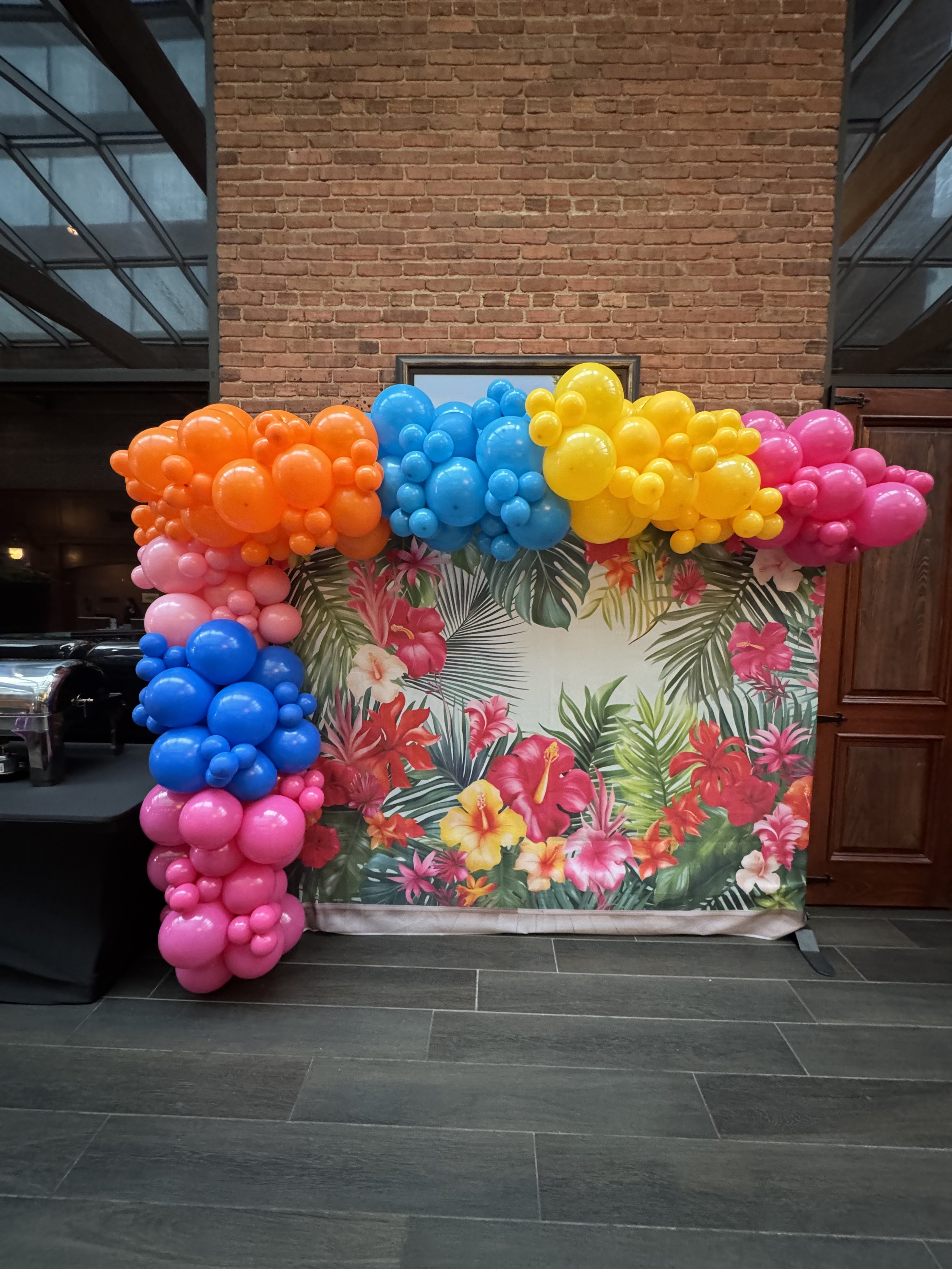 A decorative tropical floral backdrop with a colorful balloon arch featuring pink, blue, yellow, and orange balloons, in front of a brick wall inside a building.