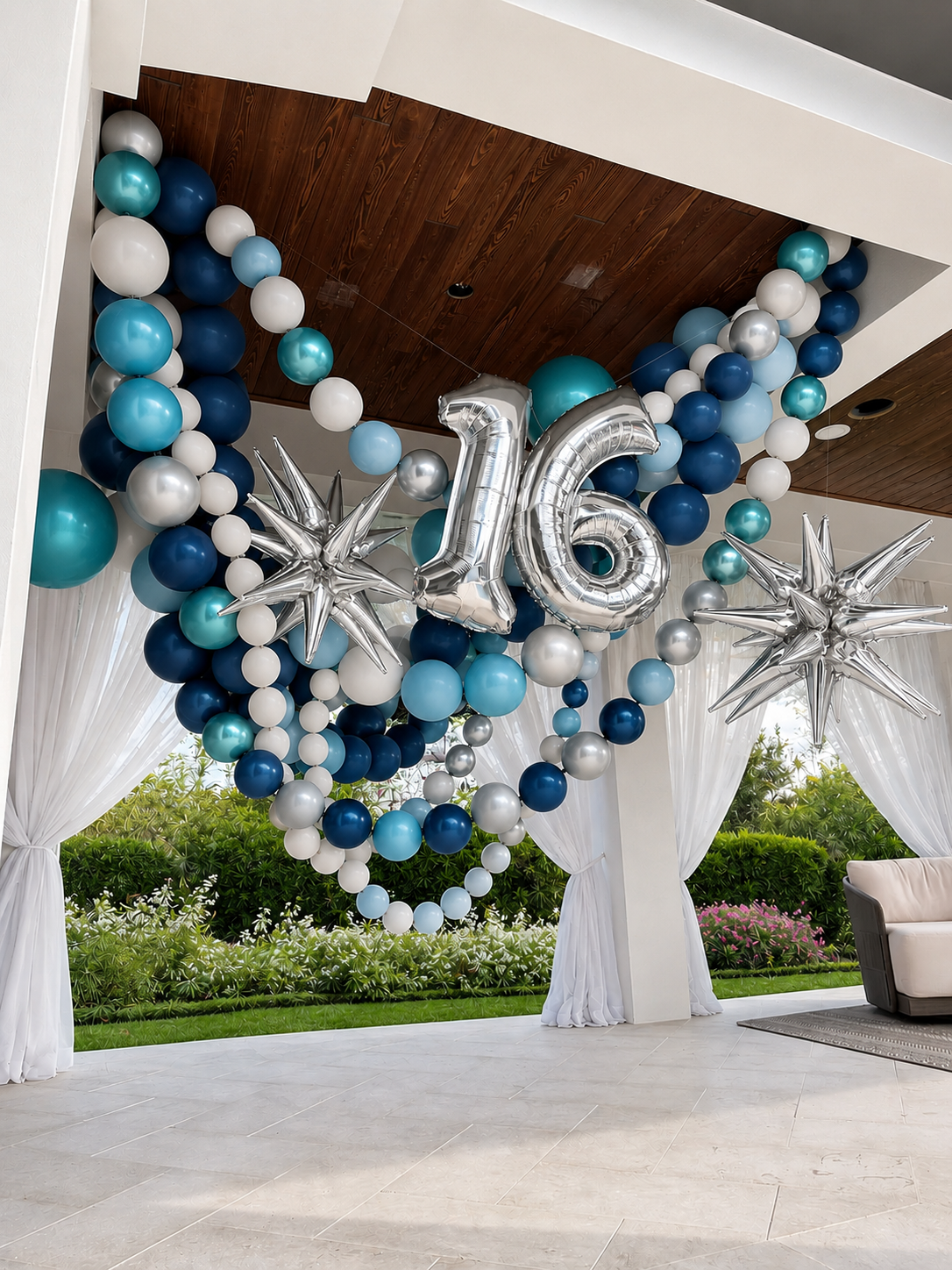 Party decoration with blue, white, silver balloons forming a heart and the number 16, with metallic star-shaped balloons, set indoors with white curtains and outdoor greenery visible.