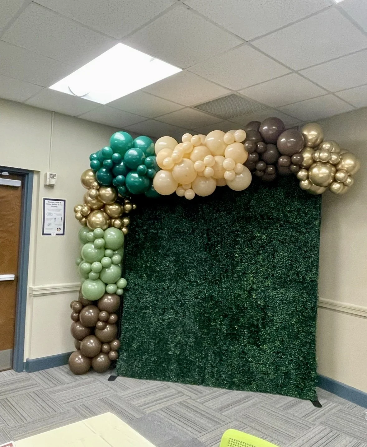 A balloon arch with green, beige, gold, and brown balloons over a dark green backdrop in an indoor room.