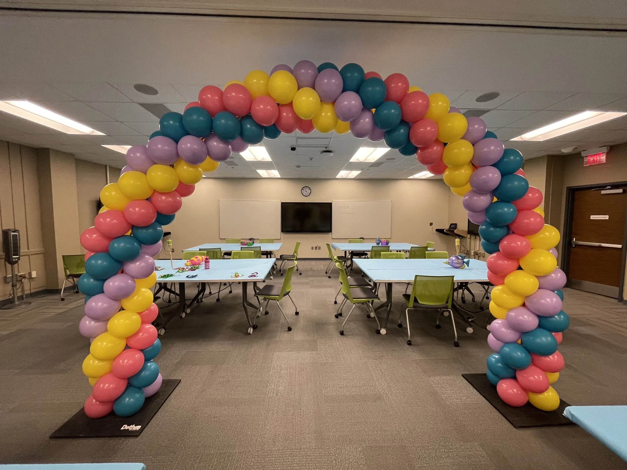 Colorful balloon arch with pink, lavender, yellow, and teal balloons in a room set up for a celebration, with tables and chairs arranged underneath.