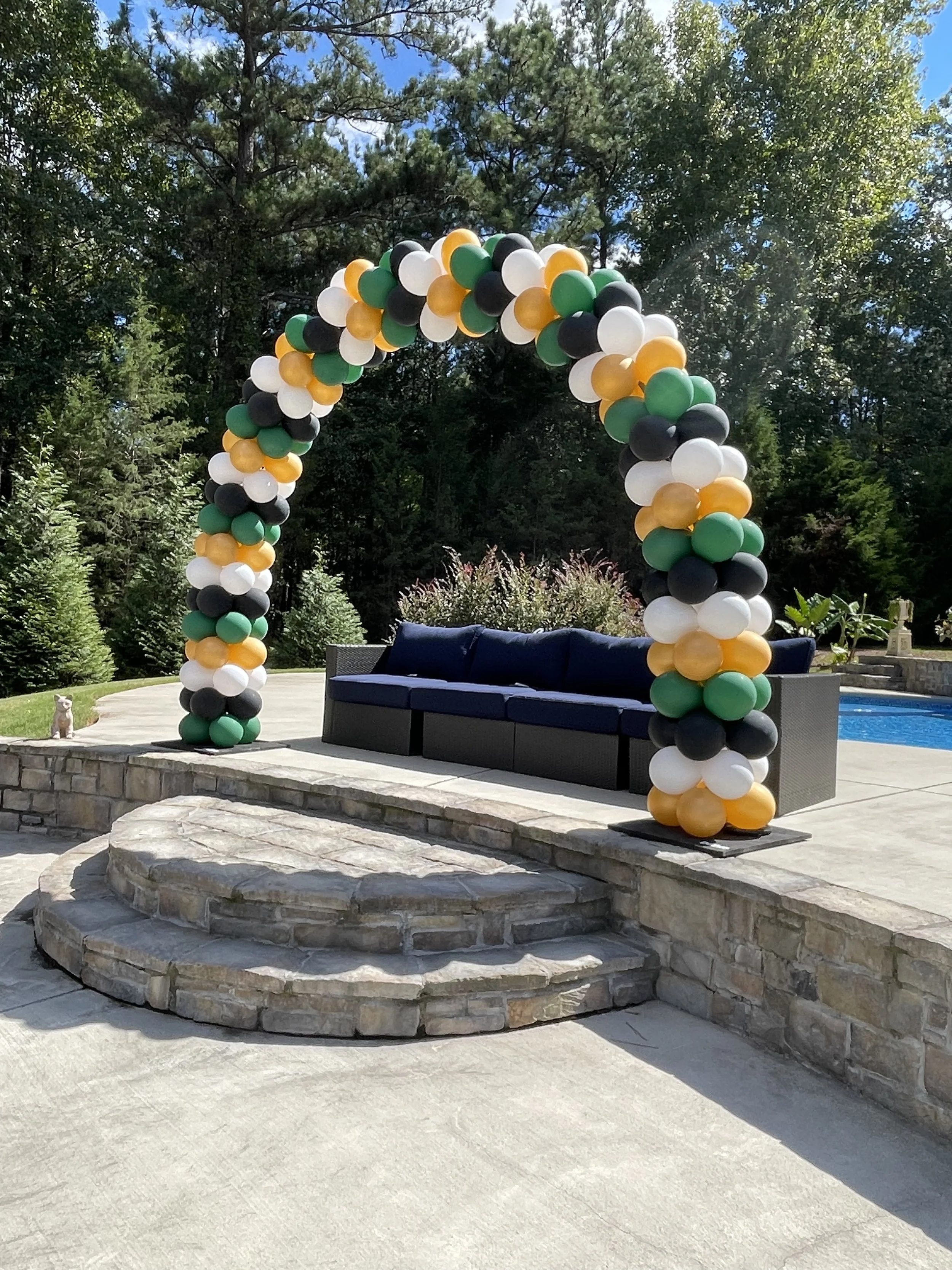 Outdoor wedding arch made of black, white, green, and gold balloons, set on a stone patio with a blue sofa underneath it. A small dog sits on the grass nearby. Tall trees and a clear blue sky are in the background.