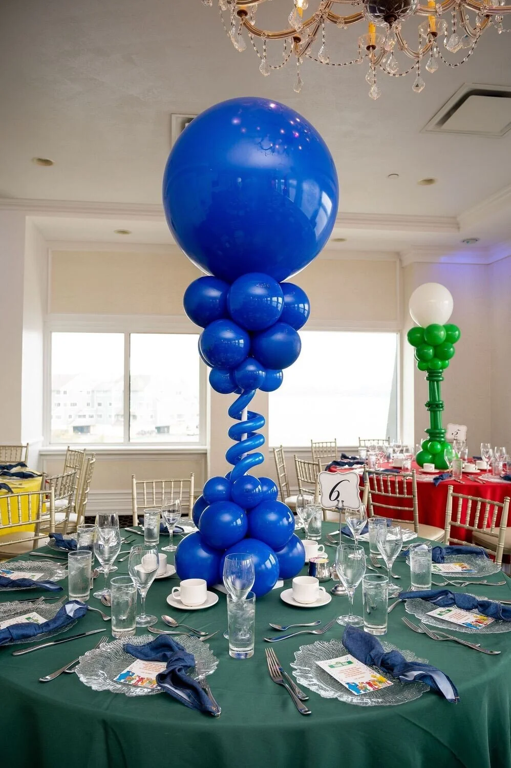 Indoor event space decorated with balloon centerpieces on tables covered with green tablecloths, set with glassware, plates, and cutlery, featuring a large blue balloon centerpiece on the main table and a smaller white and green balloon centerpiece o