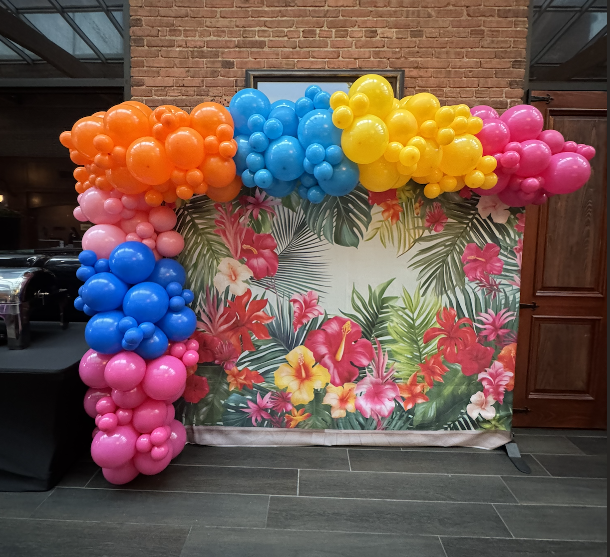 Colorful balloon arch with orange, blue, yellow, and pink balloons over a tropical floral backdrop with large green leaves and pink, yellow, and red flowers, set against a brick wall.
