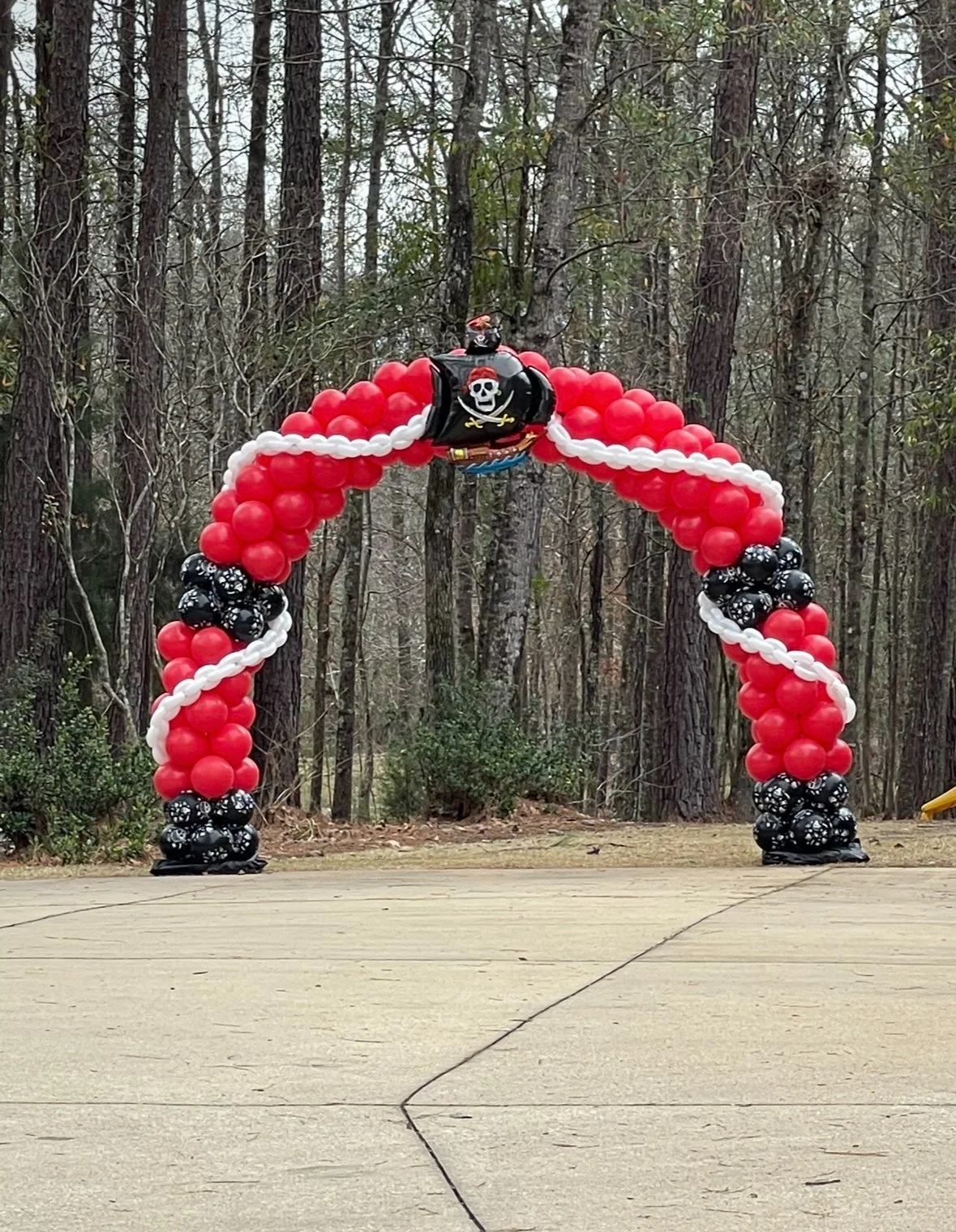 Pirate-themed balloon arch with red, black, and white balloons, featuring a pirate ship balloon at the top, set outdoors in a wooded area.