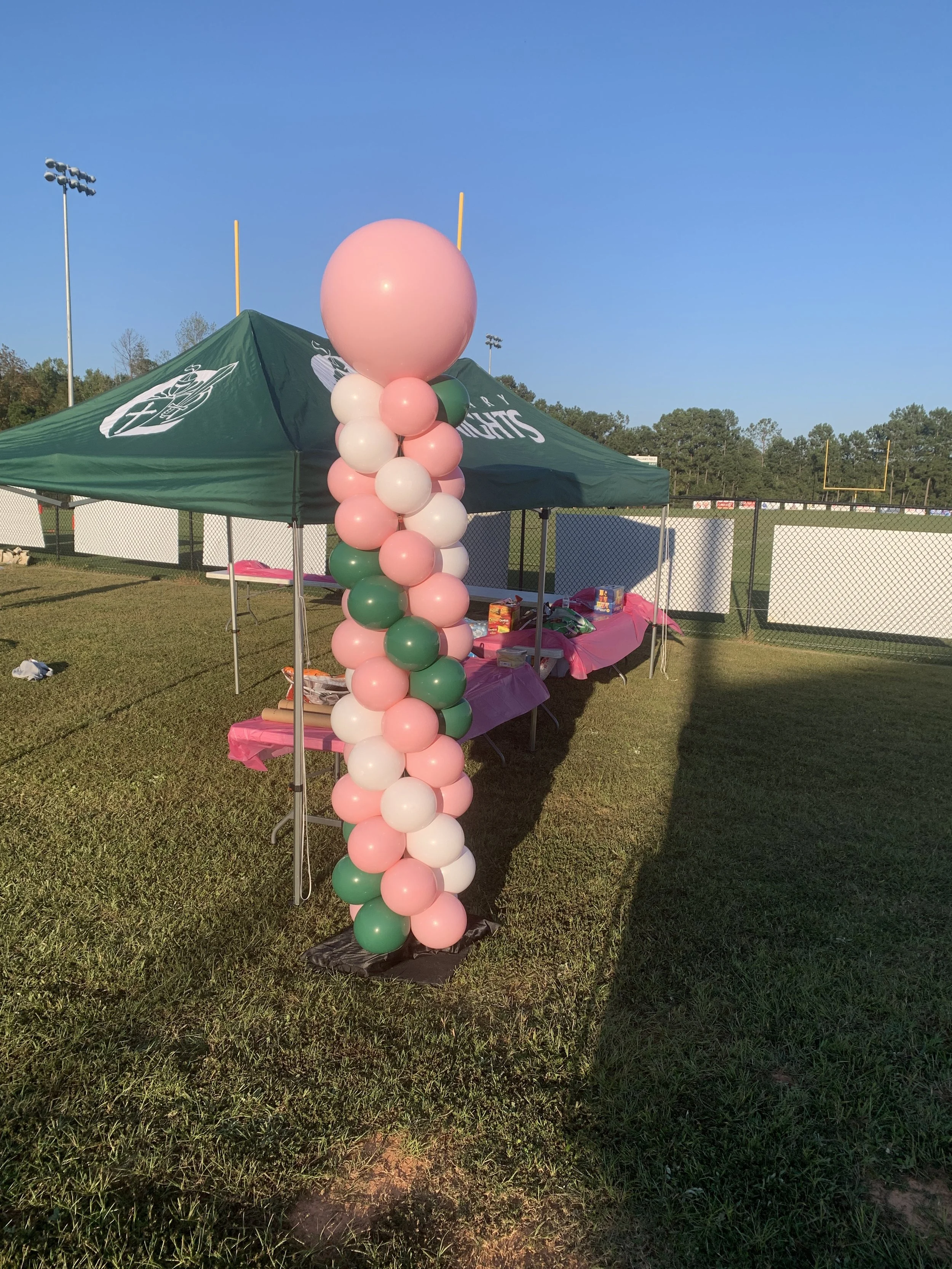 A tall balloon column with pink, white, and green balloons in front of a green sports tent at a football field. The sky is clear and the field is surrounded by a chain-link fence, with trees and stadium lights.