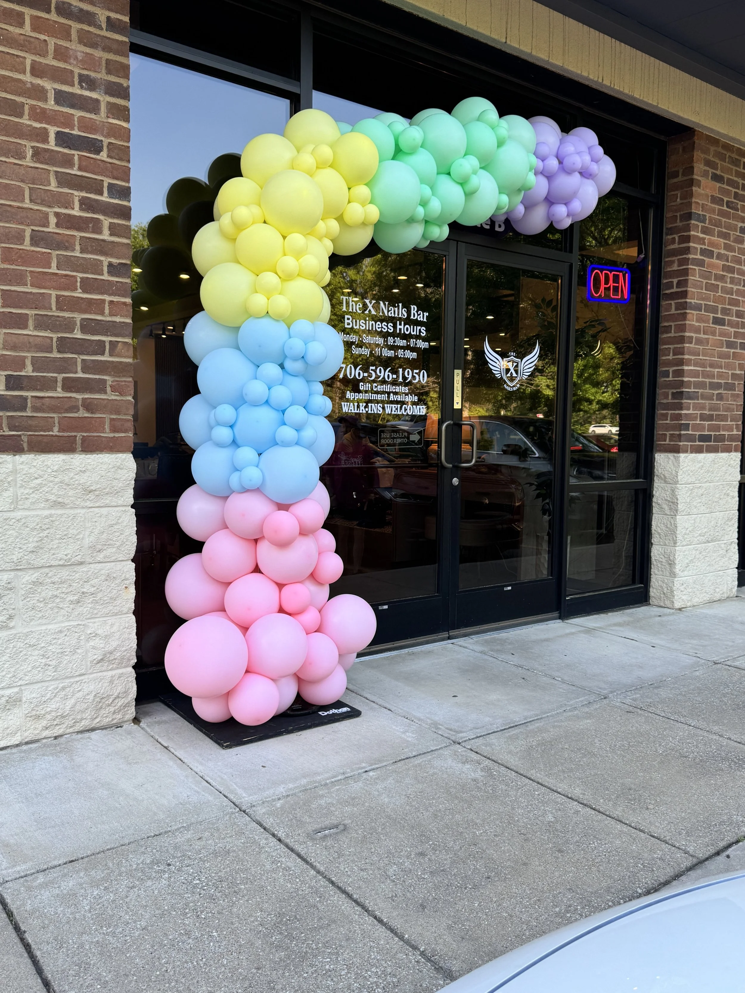 Storefront decorated with colorful balloon arch in pastel colors including yellow, blue, pink, green, purple, and black, with a neon 'Open' sign in the window of The X Nails Bar, brick and concrete sidewalk outside.
