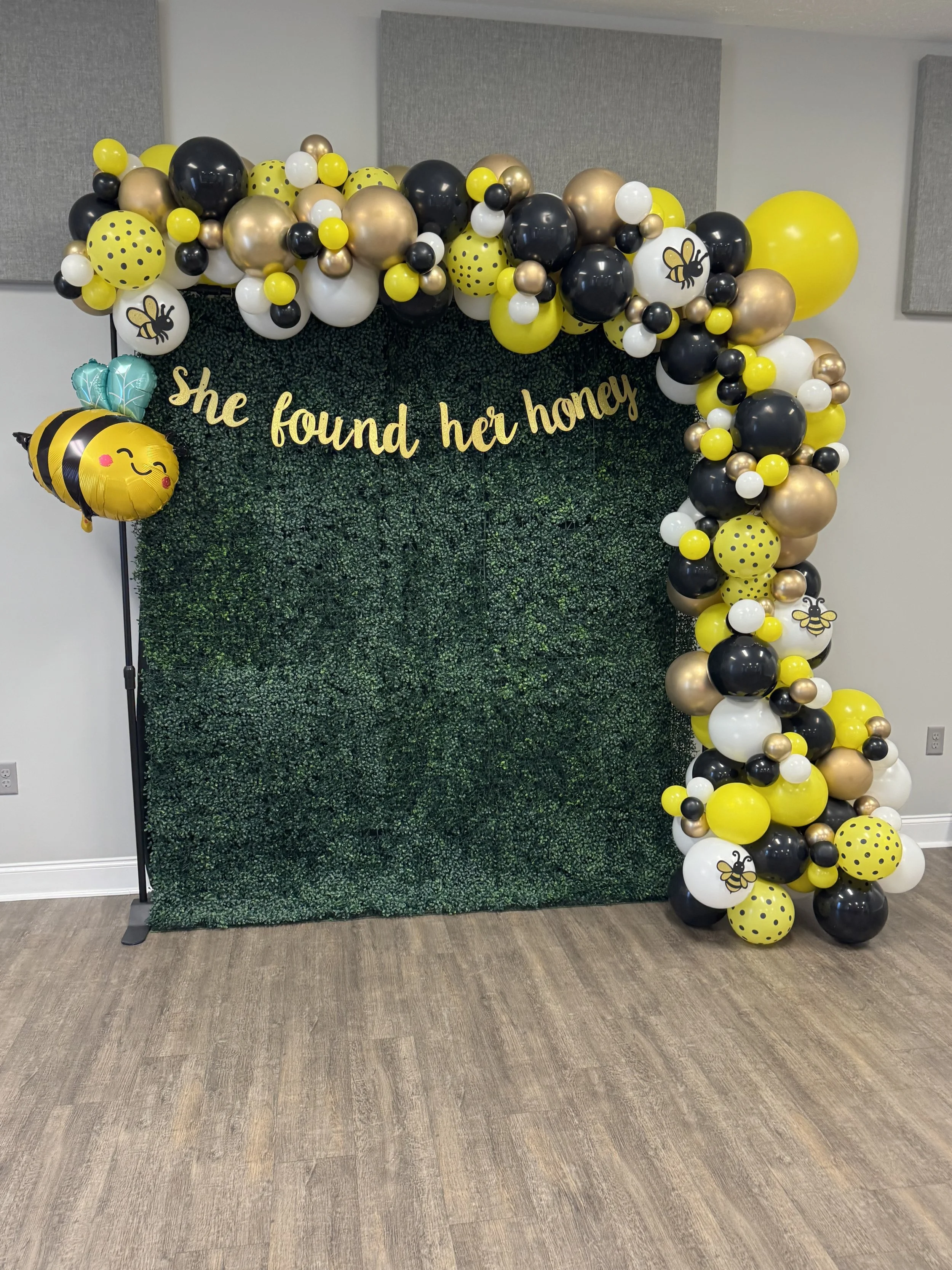 Decorative wall with a dark green hedge background and a balloon arch in black, gold, white, and yellow with bee and honeycomb designs, with the phrase "she found her honey" in yellow cursive letters.