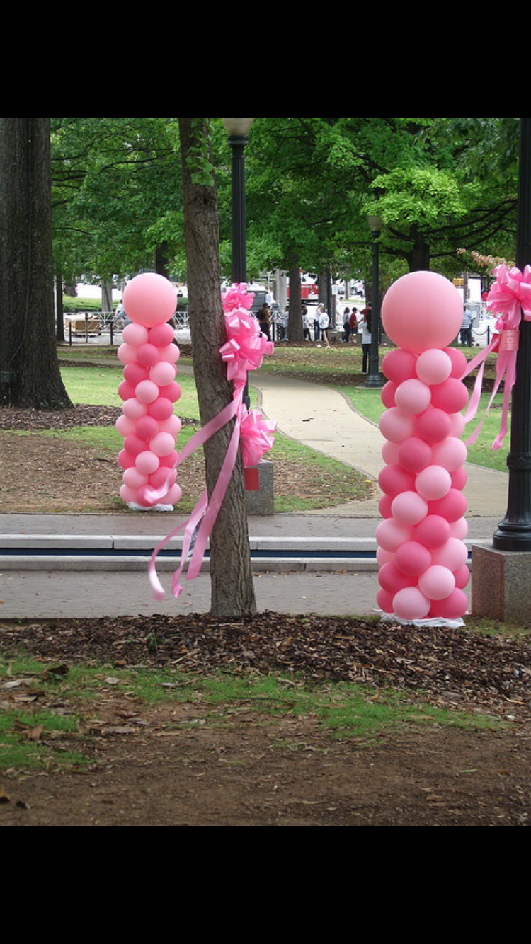 Decorative pink balloon pillars tied to trees with pink ribbons and bows in a park setting.