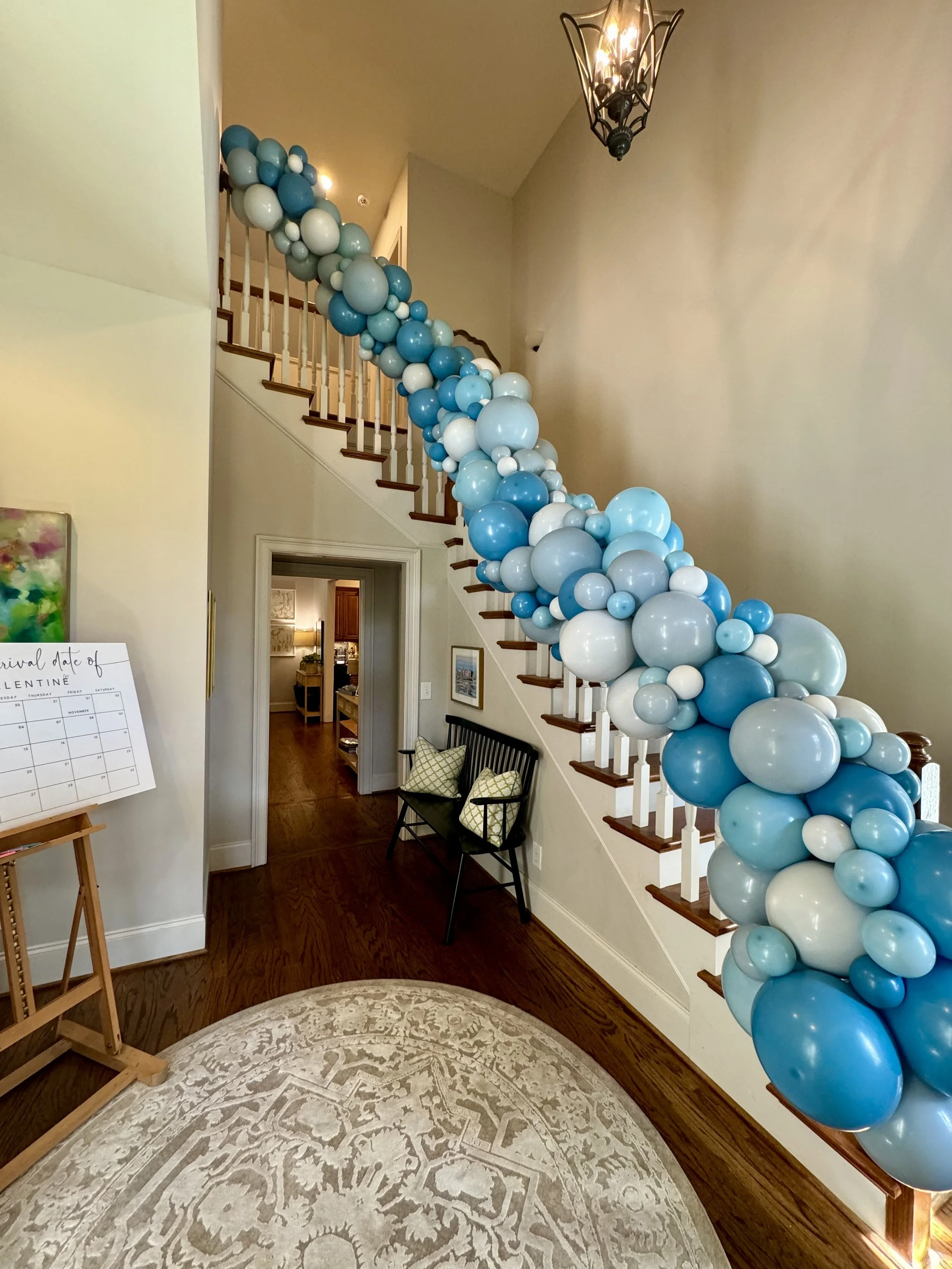 Decorative balloon arrangement in shades of blue and white along the staircase railing inside a house, with a patterned area rug on the floor and a chandelier on the ceiling.