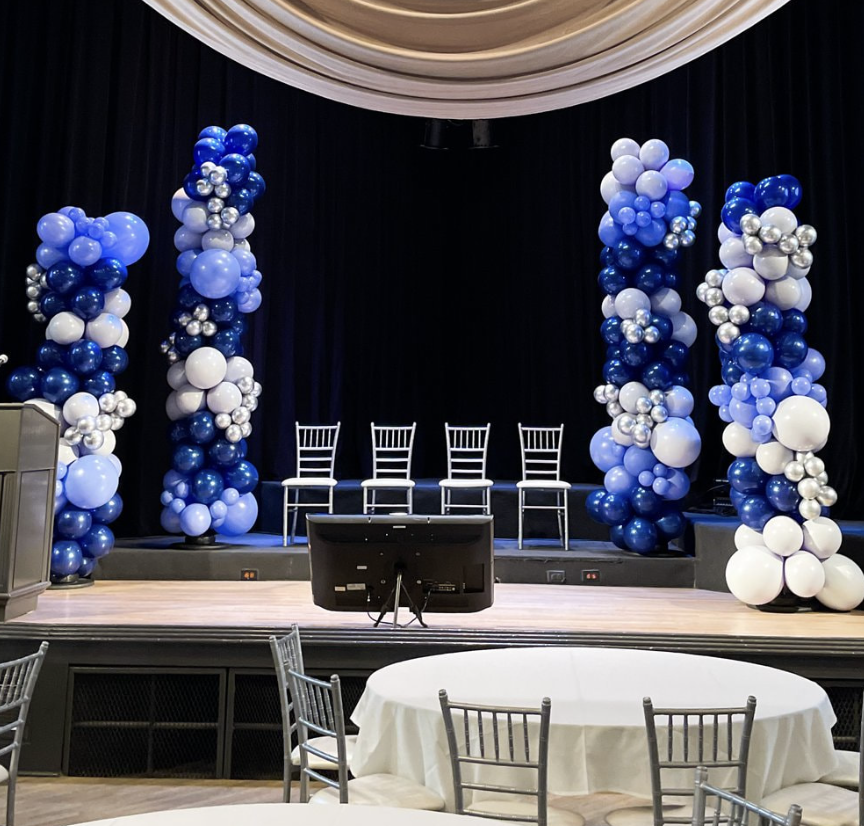 Stage decorated with tall balloon columns in shades of blue, white, and silver, set behind four white chairs, with a round table and chairs in the foreground, and a black curtain backdrop.