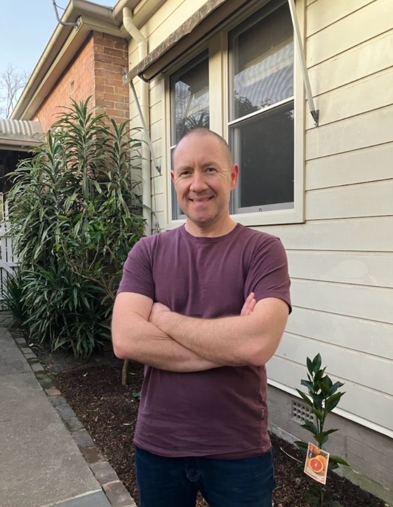 A man standing outside a house with his arms crossed, smiling, next to a small plant with a tag. The house has a beige siding exterior and a window behind him. There are shrubs and a brick wall in the background.