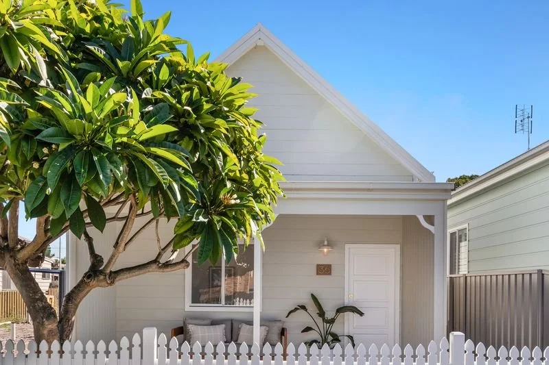White house with a sloped roof, small front porch with seating, and a large green tree in front.