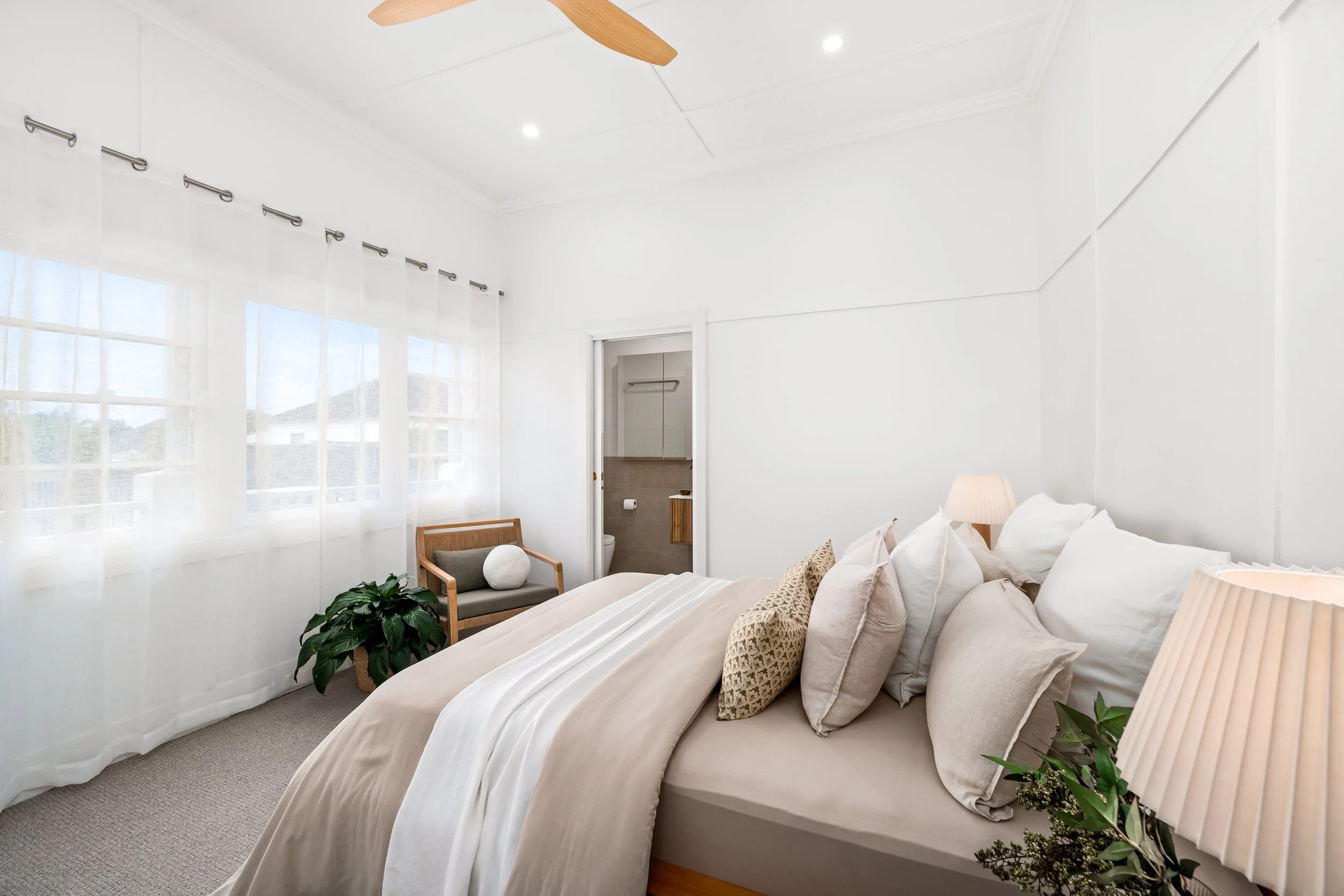 Bright bedroom with white walls, large windows with sheer curtains, a bed with beige bedding and multiple pillows, a wooden armchair with cushions, potted plants, and a view into an en-suite bathroom with a toilet.
