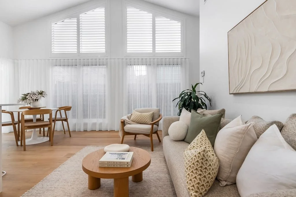 Bright living room with white walls, large windows with white shutters and sheer curtains, a beige sofa with multiple pillows, a round wooden coffee table with books, a beige armchair with a pillow, and a dining table with chairs in the background.