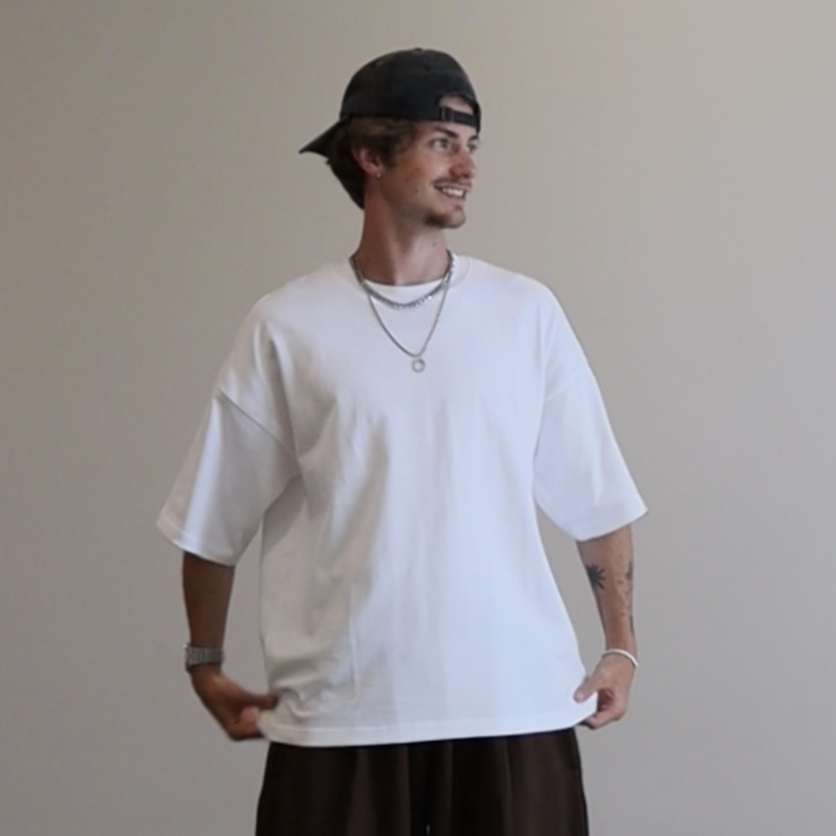 A young man wearing a black backwards cap, white oversized T-shirt, layered necklaces, and brown pants, standing against a plain wall, smiling and looking to his left.