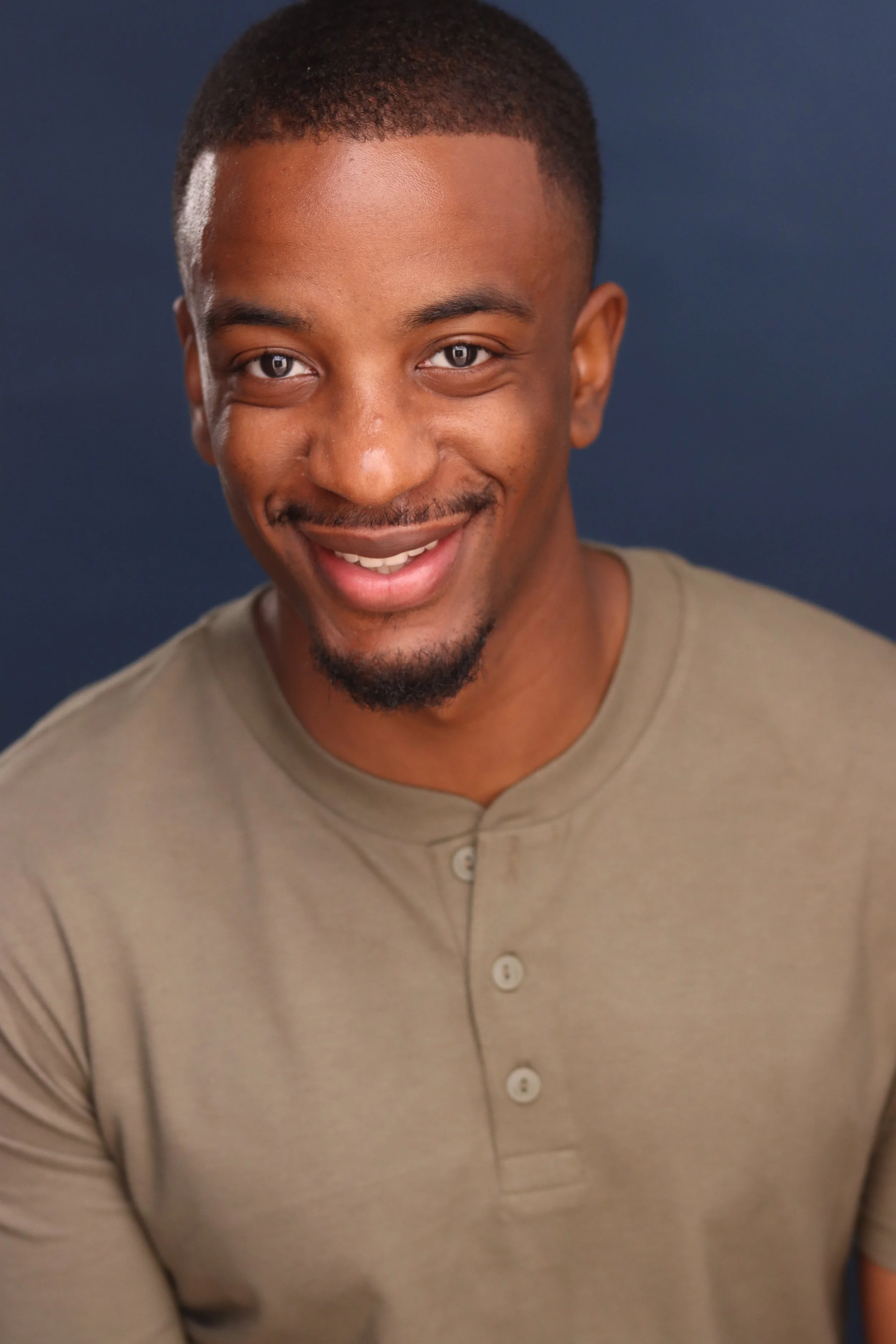 close-up of a smiling young man with short hair, goatee, and wearing a beige button-up shirt, against a dark blue background.