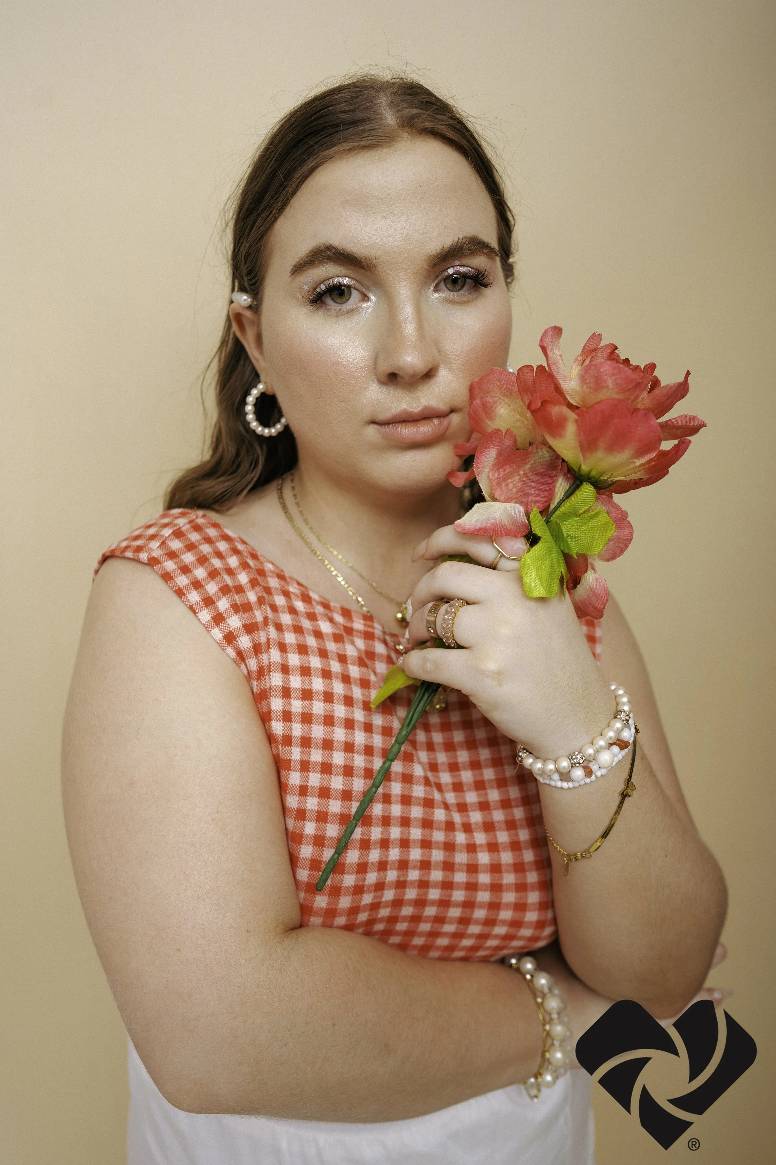 A woman with light skin and brown hair holding a pink and yellow flower. She is wearing pearl earrings, a gold chain necklace, bracelets, multiple rings, and a red gingham sleeveless top. The background is plain beige.
