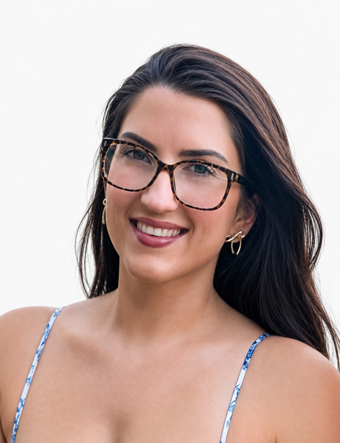 Close-up of a smiling woman with long dark hair, wearing tortoise shell glasses, gold earrings, and a blue patterned spaghetti strap top.
