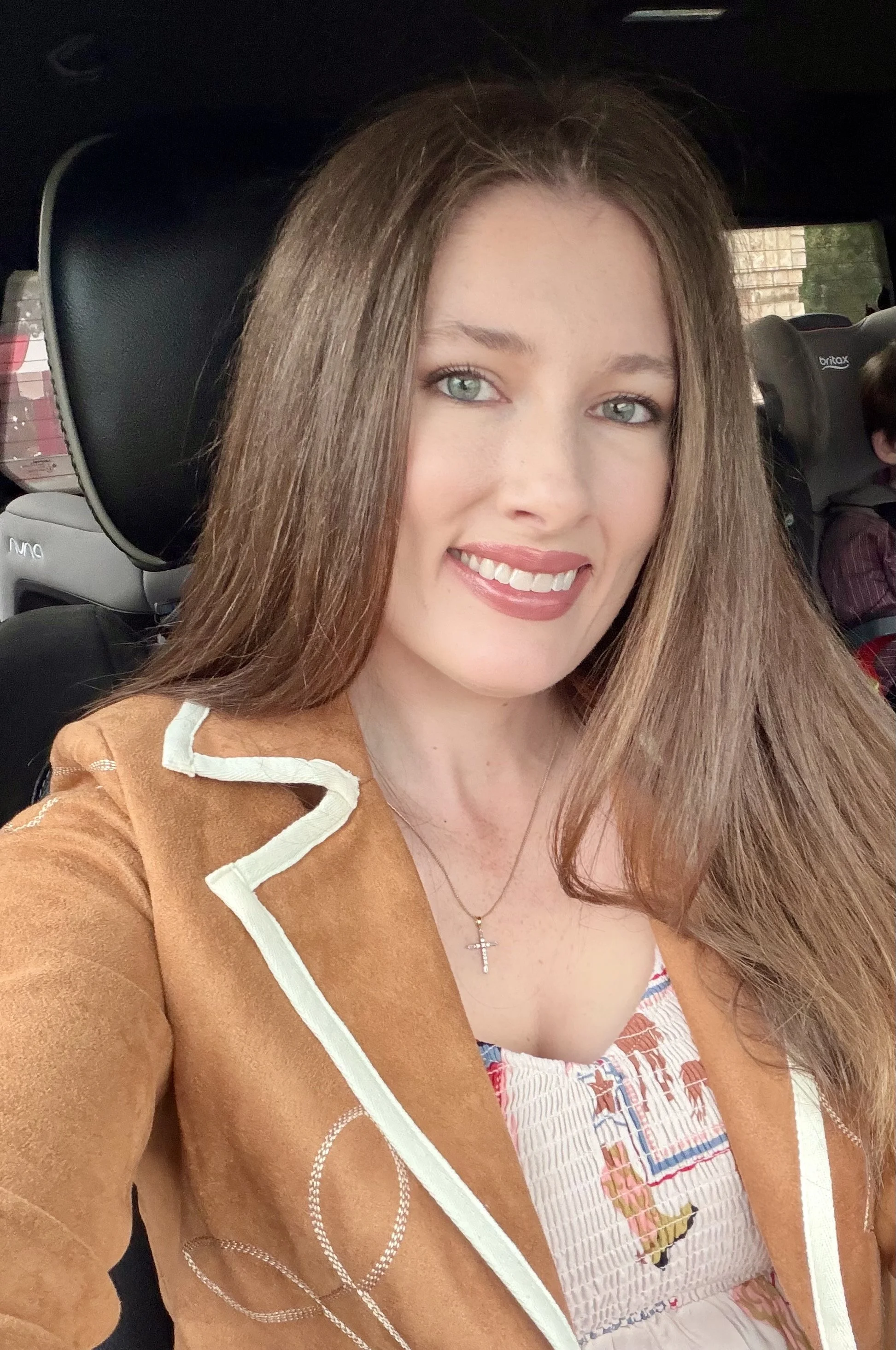 A young woman with long brown hair, blue eyes, and wearing a tan jacket with white trim. She is smiling and wearing a cross necklace. In the background, a child is seated in a car seat.