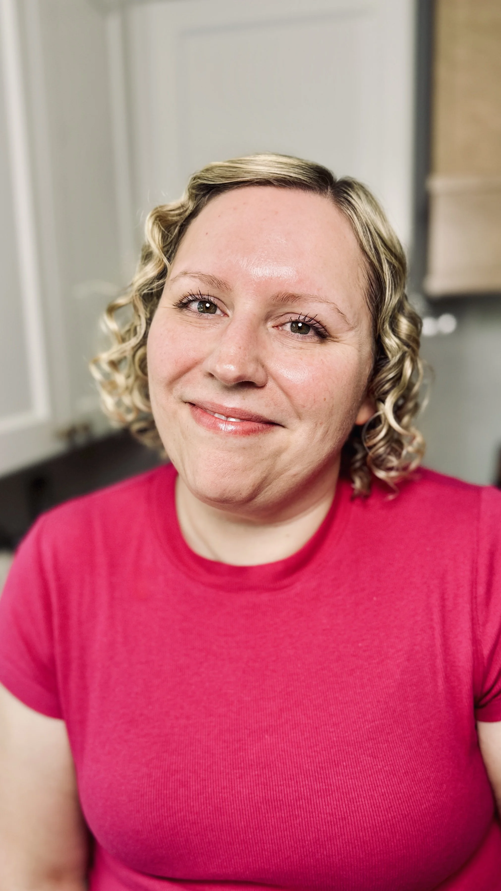 A woman with curly blonde hair smiling, wearing a bright pink shirt, in an indoor setting.