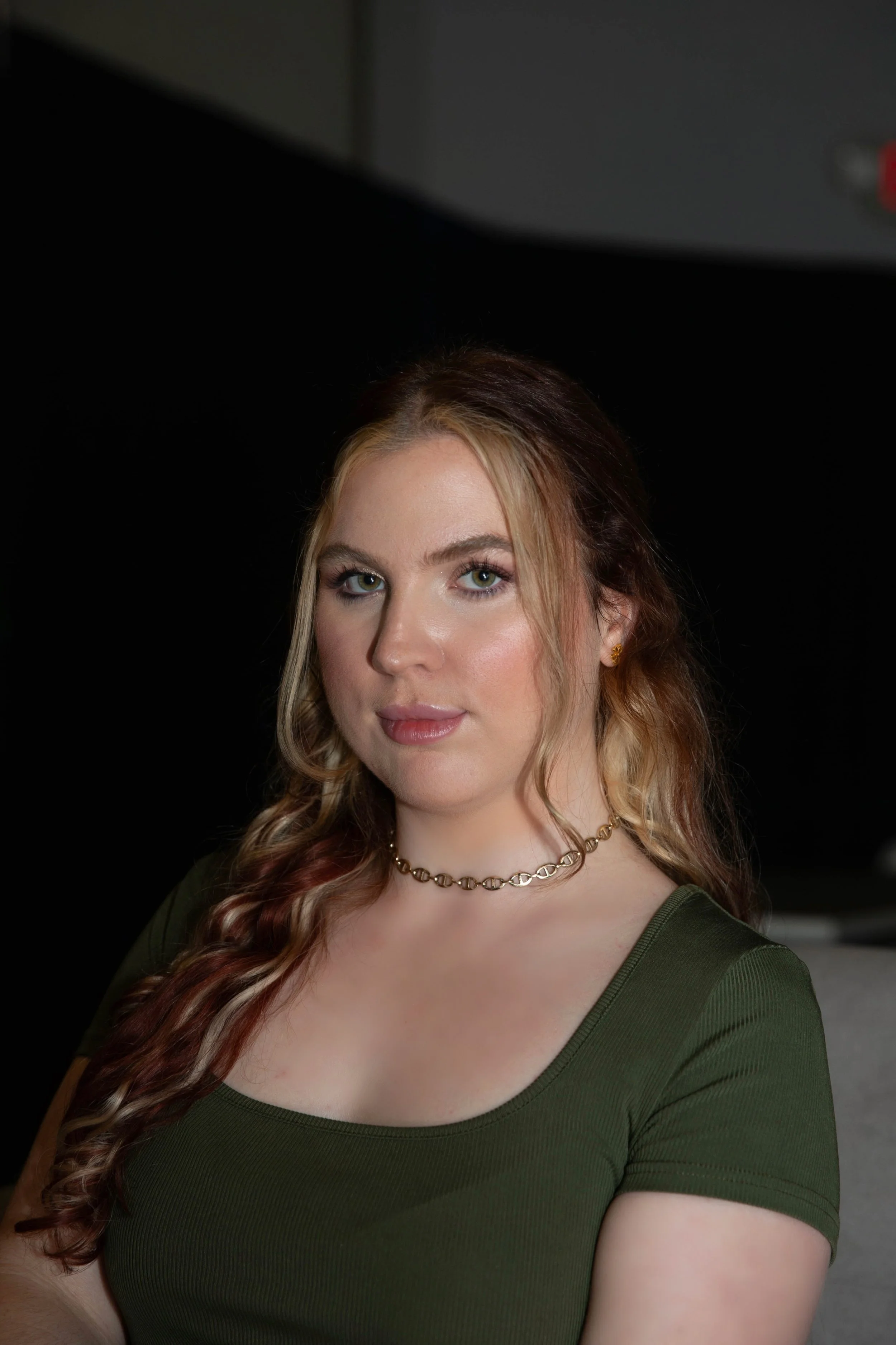 Close-up of a woman with long, wavy hair, wearing a green top and a gold chain necklace, against a dark background.