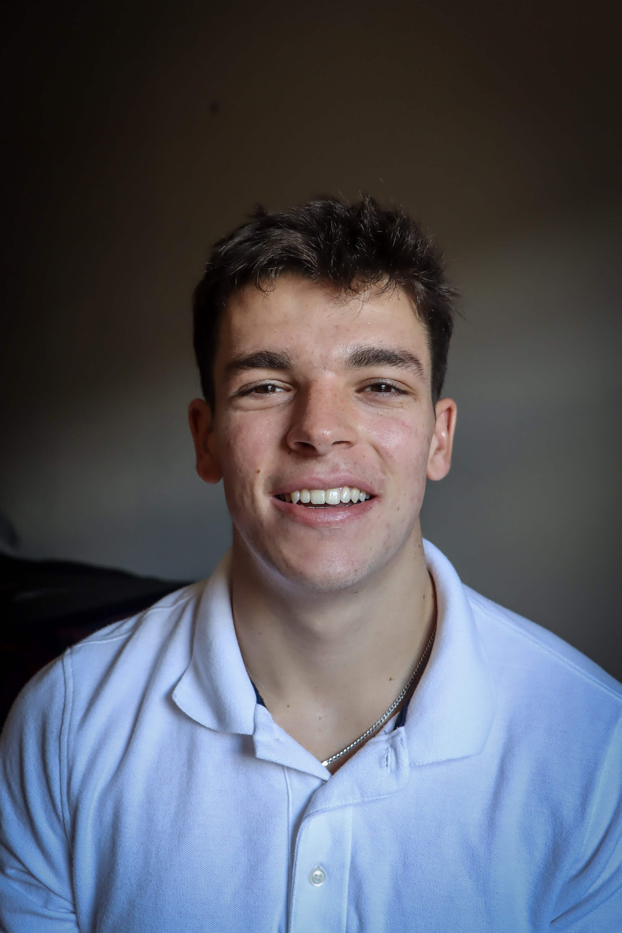 A young man with short dark hair, smiling, wearing a light blue collared shirt with a chain necklace.