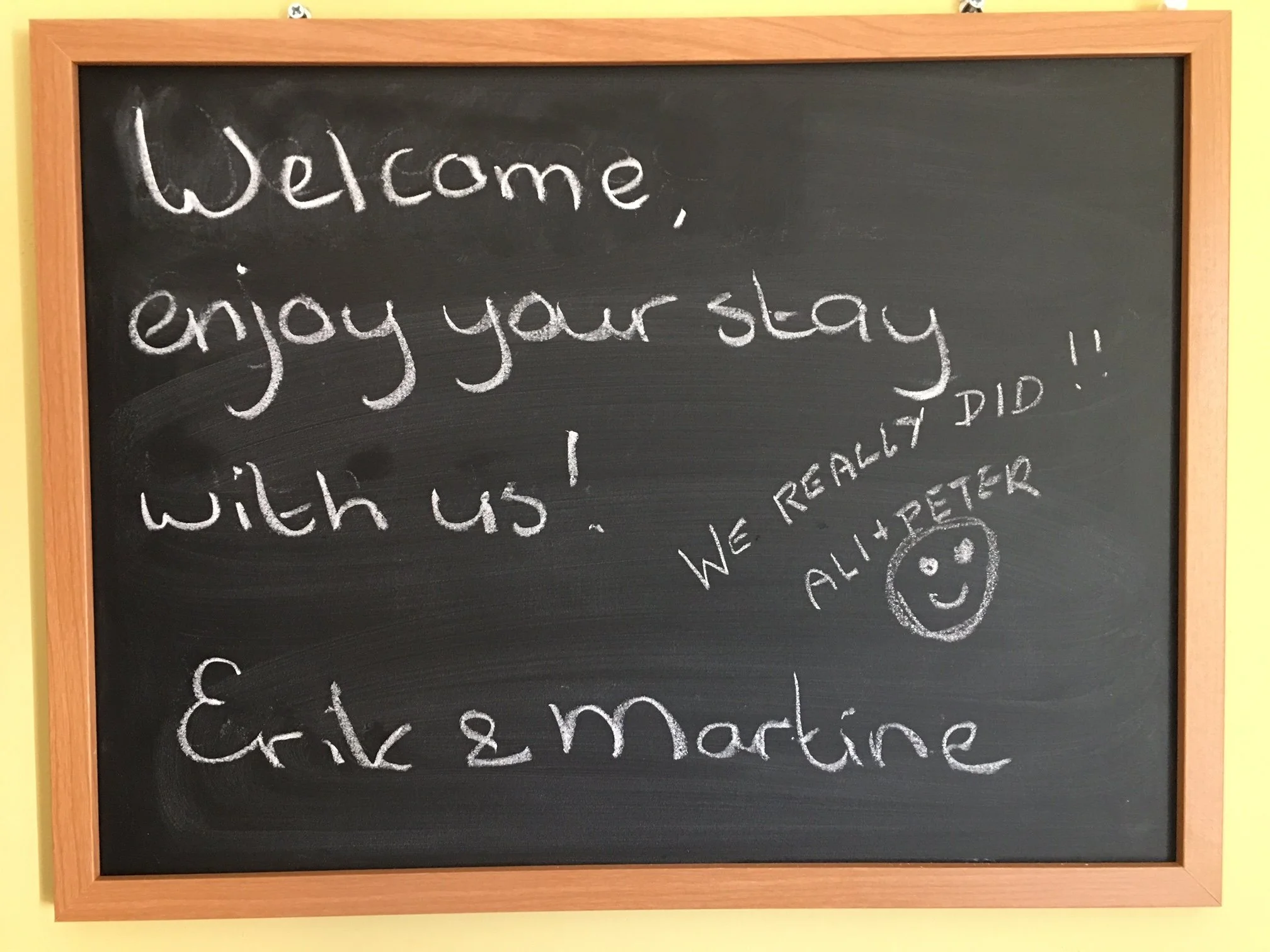 A blackboard with a wooden frame displaying the message 'Welcome, enjoy your stay' written in white chalk, with additional handwritten notes and a smiley face drawing.