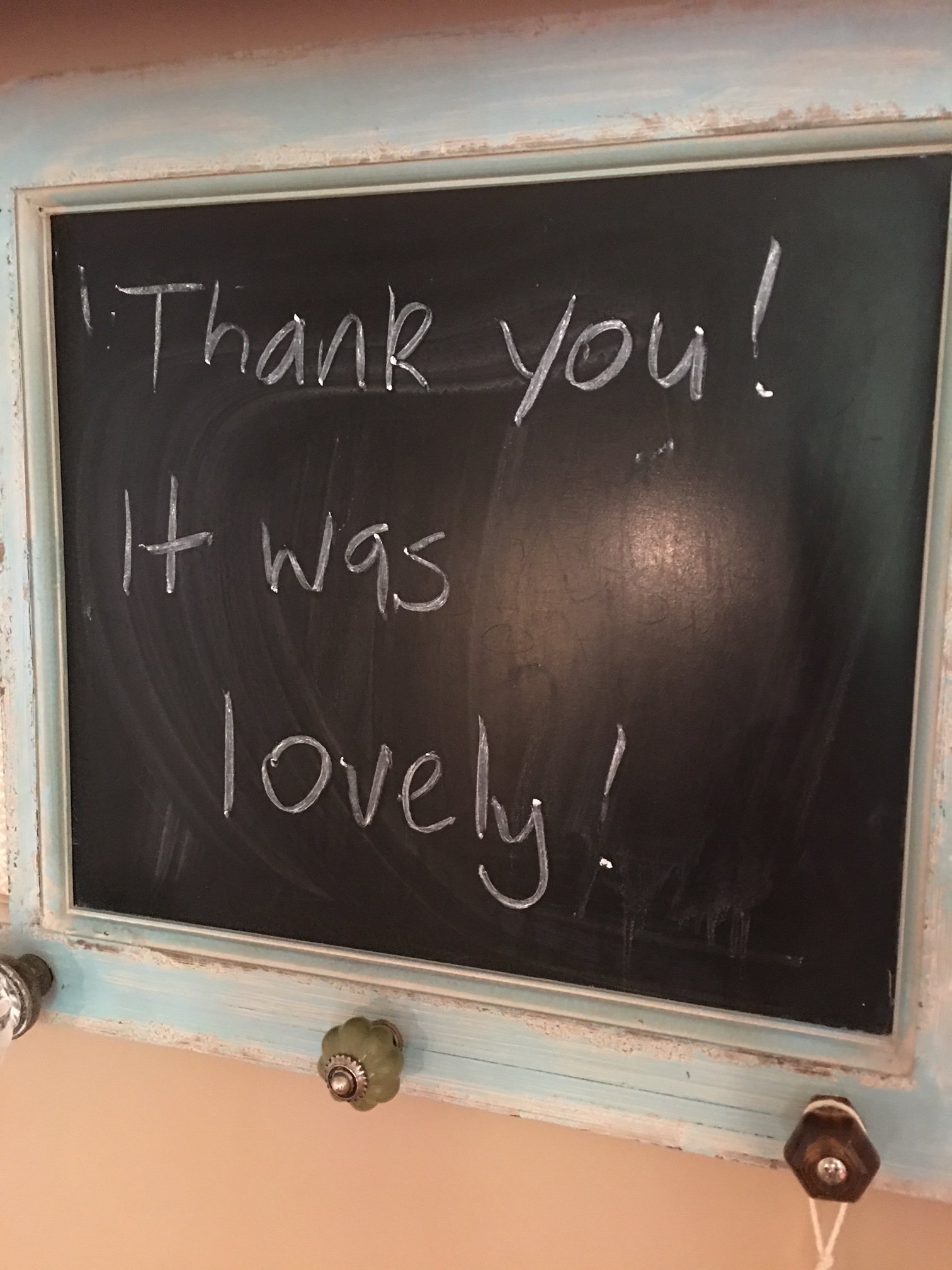 A black chalkboard with a white frame, showing handwritten words 'Thank you! It was lovely.' in white chalk, placed on a wall with a beige background.