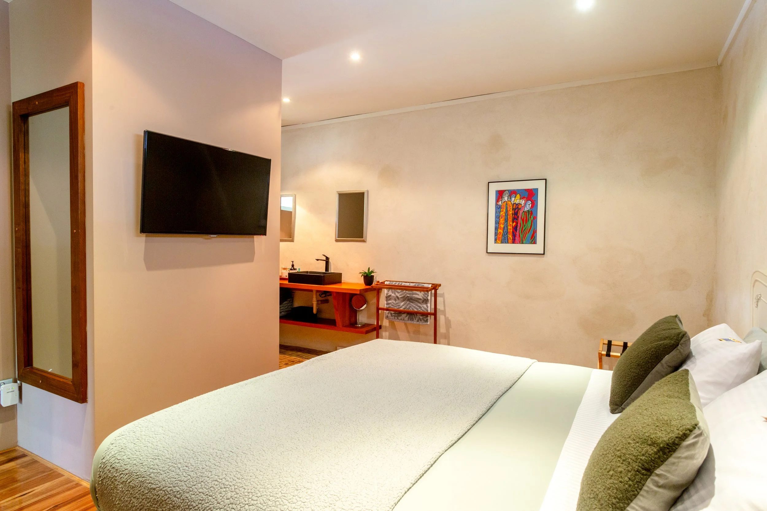 Hotel room with a bed, pillows, painting, wall-mounted TV, and a small wooden table with a sink and mirror in the background.