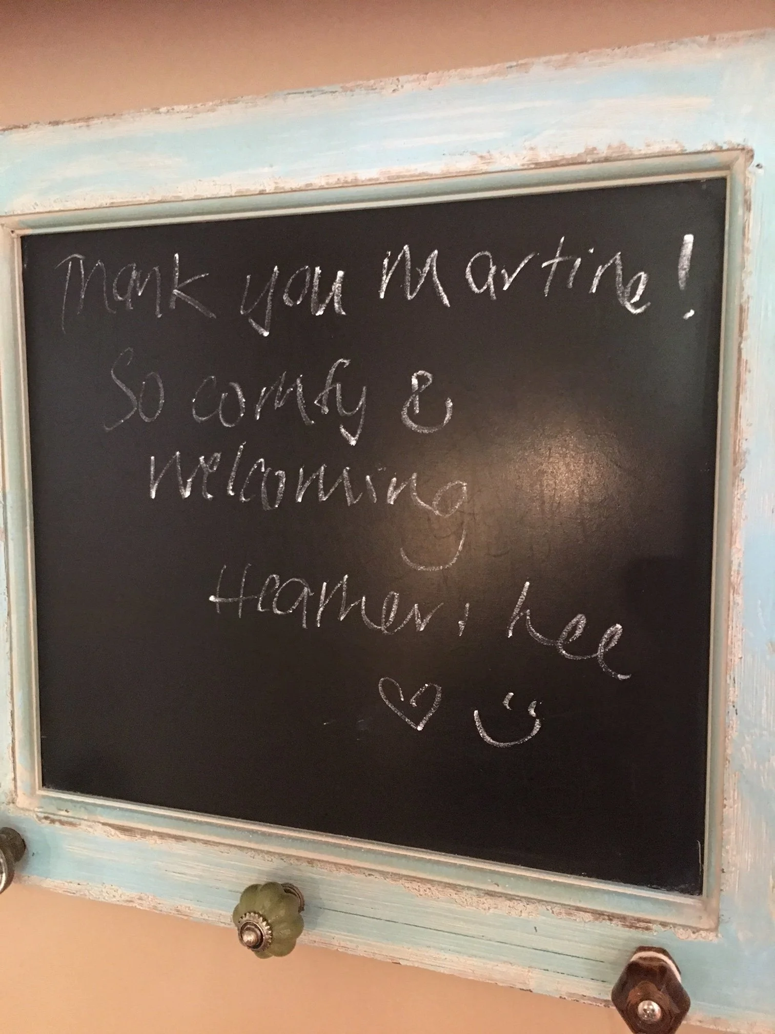 A black chalkboard with a light blue wooden frame. Handwritten in white chalk on the board is the message: "Thank you Martin! So sorry & Welcoming." A small heart drawing and a smiley face are also visible on the board.