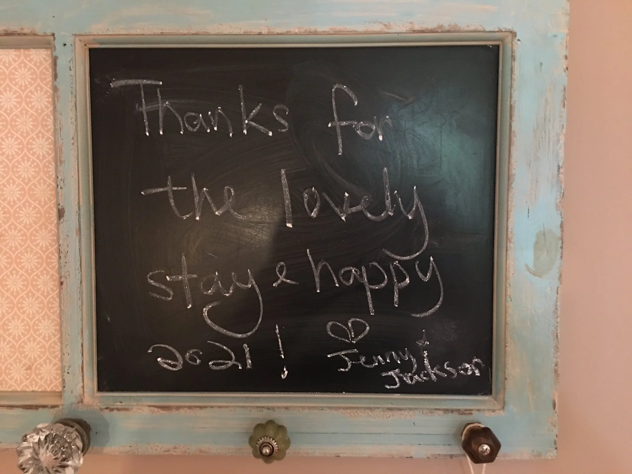 A black chalkboard with a wooden frame displaying the message 'Thanks for the lovely stay & happy Halloween' written in white chalk. The surrounding wall shows signs of peeling paint and an adjacent patterned wallpaper on the left.