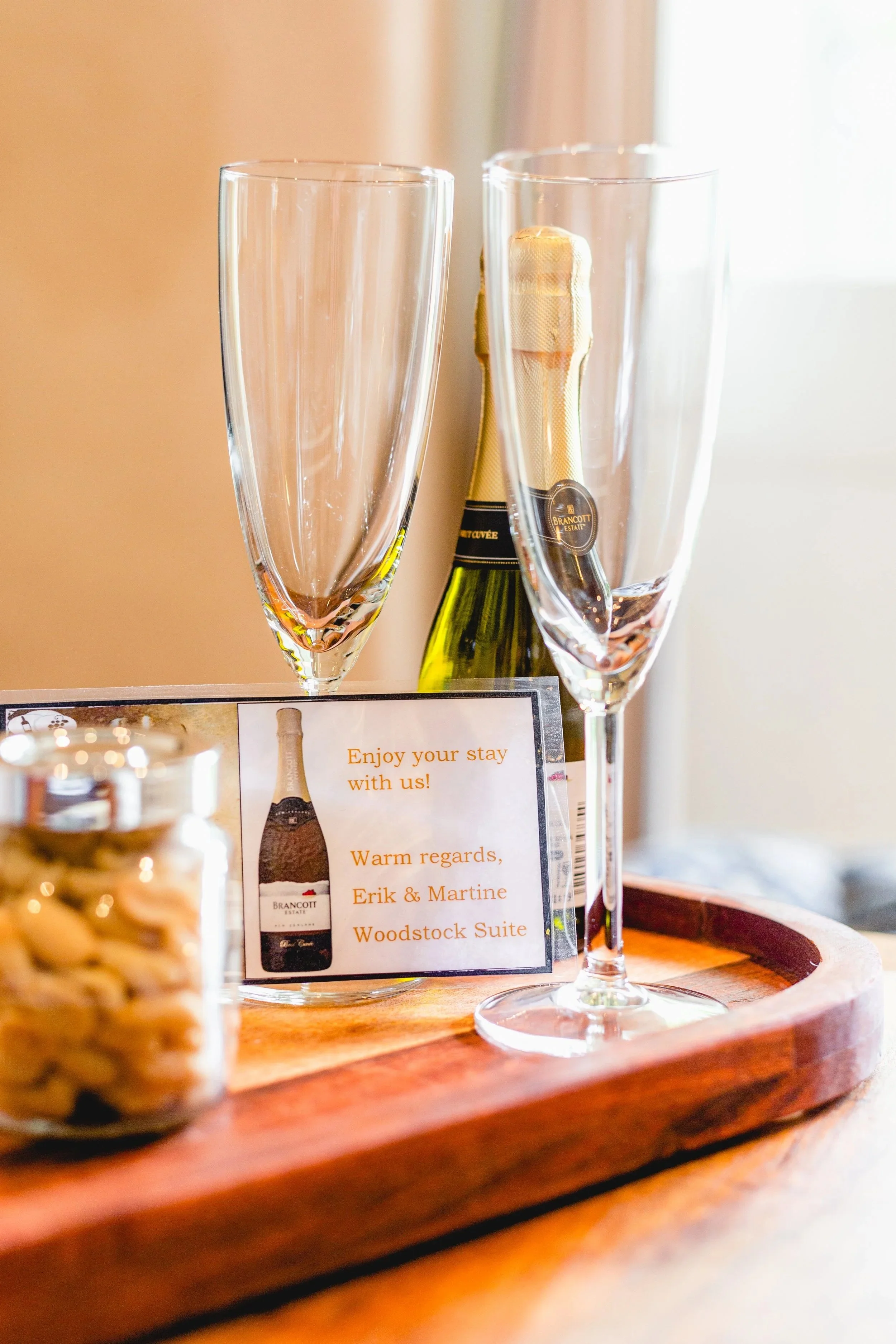 Two empty champagne flutes behind a bottle of Brancott Estate wine, one of the treats to welcome you at the Woodstock Suite. The glasses are on a wooden tray and the background is brightly lit with natural light.