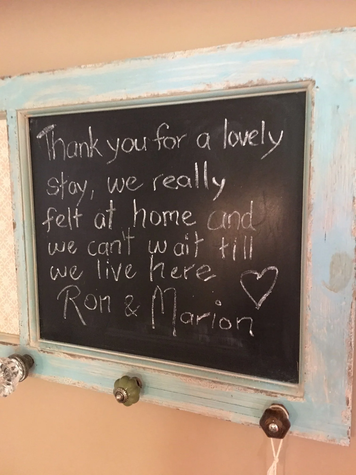 A black chalkboard with a heart drawn in the lower right corner. The board has a weathered light blue wooden frame. There are visible wall and ceiling in the background, and some indistinct objects at the bottom of the frame.