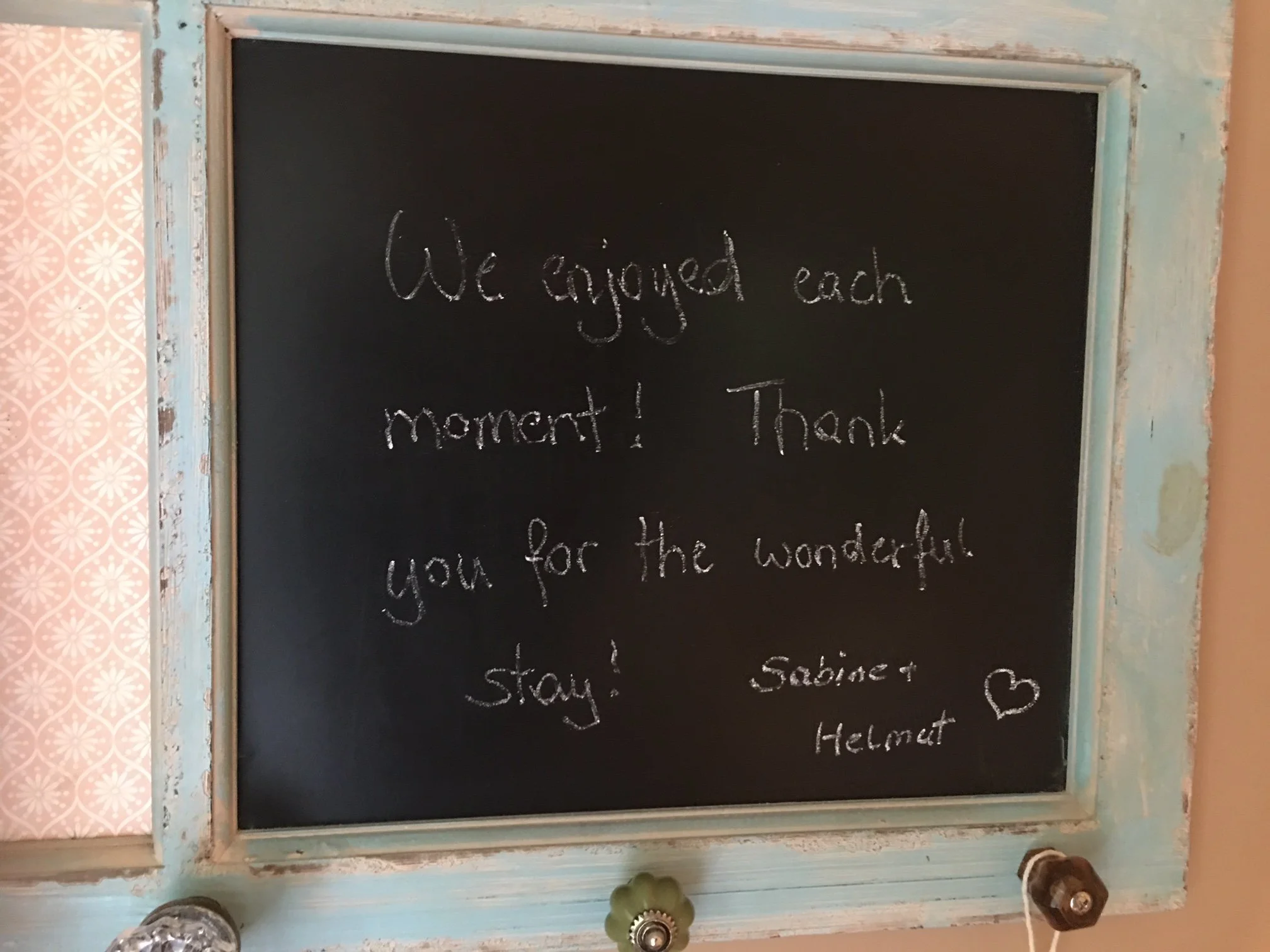 A chalkboard with a message written in white chalk reading: 'We enjoyed each moment! Thank you for the wonderful stay!' The chalkboard is framed with a weathered, light blue border, and part of a pink patterned wallpaper is visible on the left side o