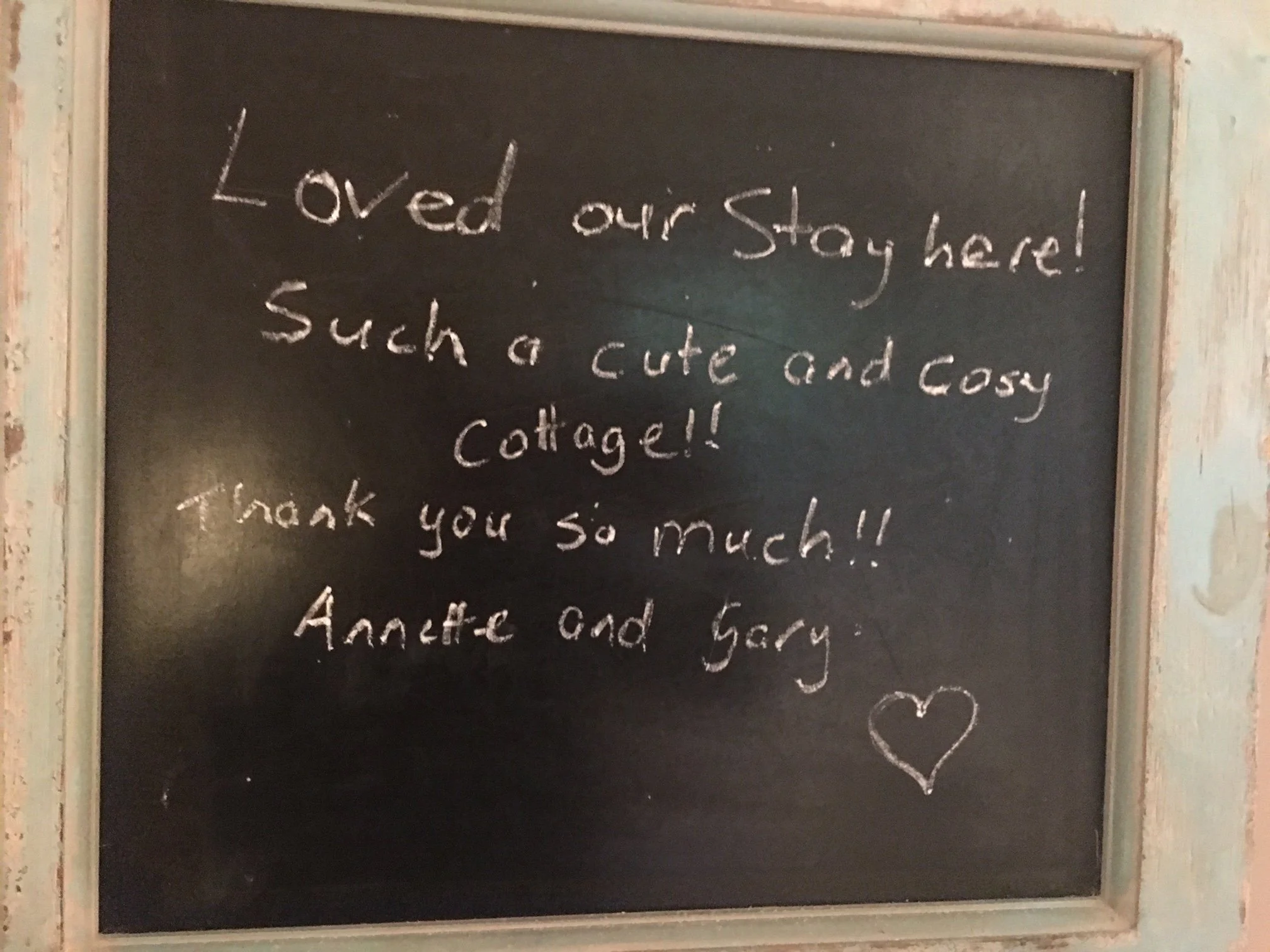 A chalkboard with handwritten message reading, "Loved our stay here! Such a cute and cozy cottage!!" and a drawn heart.