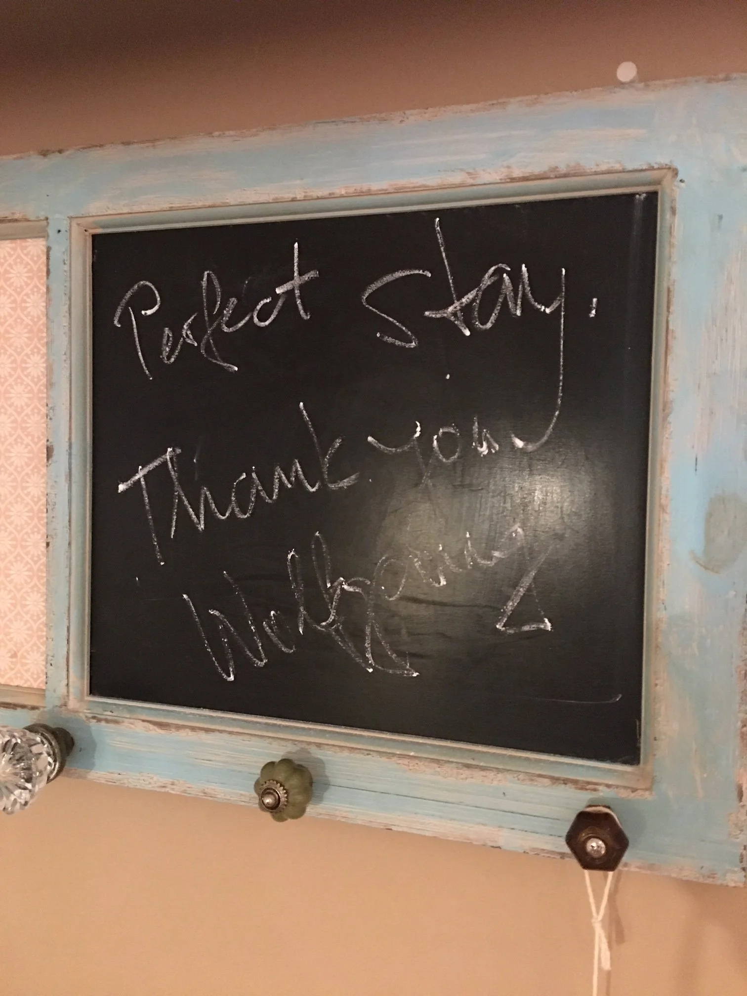 A chalkboard with the words 'Perfect Stay! Thank you!' written on it, hung on a wall with a distressed frame and knobs at the bottom.