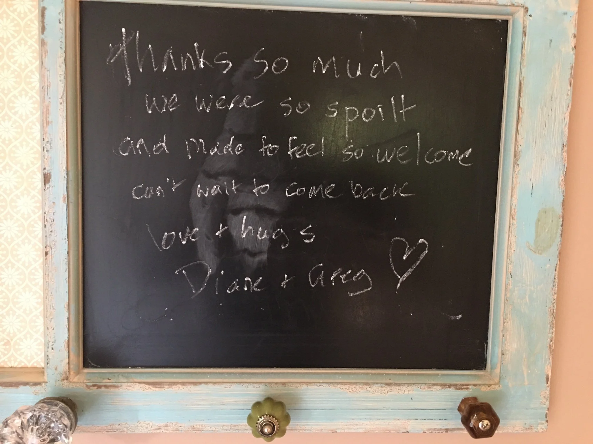 A black chalkboard with a wooden frame, containing handwritten text that reads: 'Thanks so much. We were so spoiled and made to feel so welcome. Can't wait to come back. Love & hugs, Diana'.