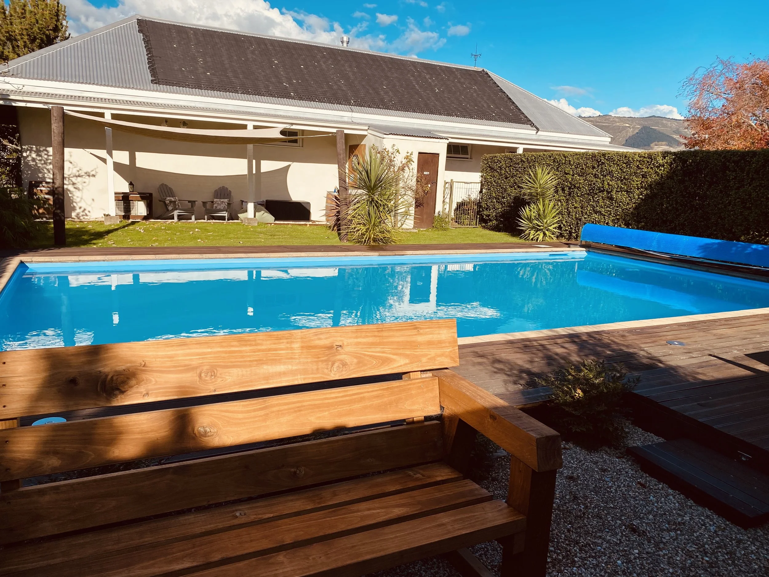 View of the private garden with solar heated swimming pool, wooden deck and outdoor seating area with chairs and plants, under a blue sky with clouds.