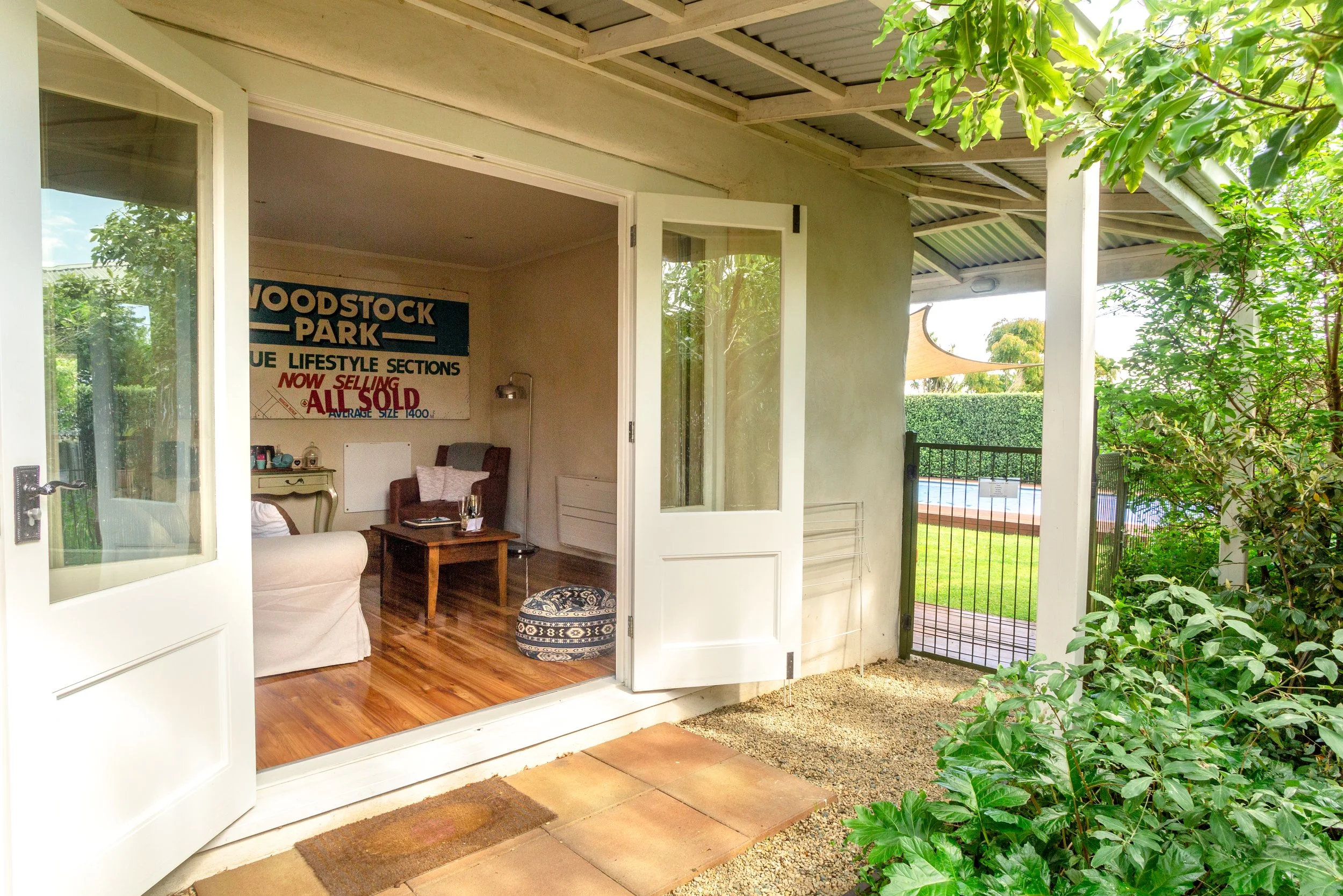 Open glass door revealing a living room with wooden floors, a sofa, a small table, and a vintage-style poster on the wall. Outside, there is a fenced yard with greenery and a swimming pool.