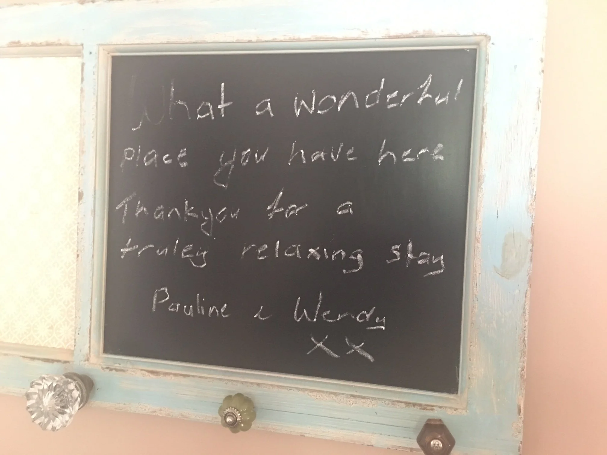 Decorative chalkboard with handwritten message, decorated with glass gems at the corners, framed with distressed light blue wood, mounted on a wall with pale pink and cream wallpaper.