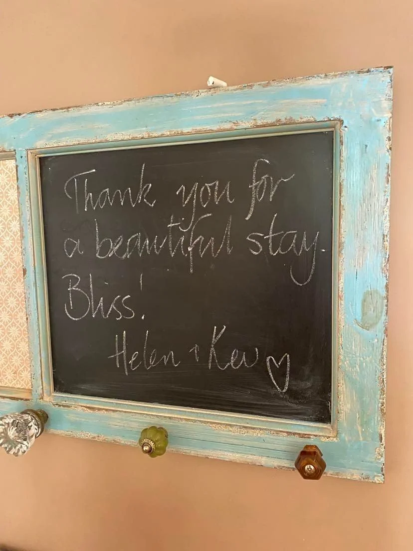 A decorative chalkboard with a distressed turquoise frame hangs on a pink wall. The chalkboard has a handwritten message that says, "Thank you for a beautiful stay. Bliss. Helen & Kay ❤️".