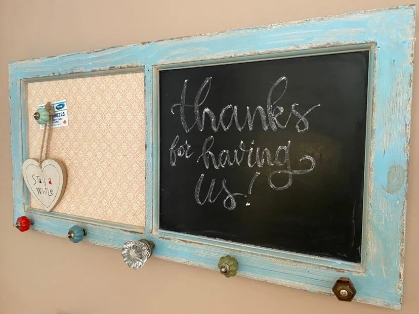A decorative wall board with a chalkboard and corkboard section, surrounded by a distressed light blue frame, with colorful knobs at the bottom and a hanging heart-shaped ornament that says 'Stay a While'. The chalkboard displays 'thanks for having u